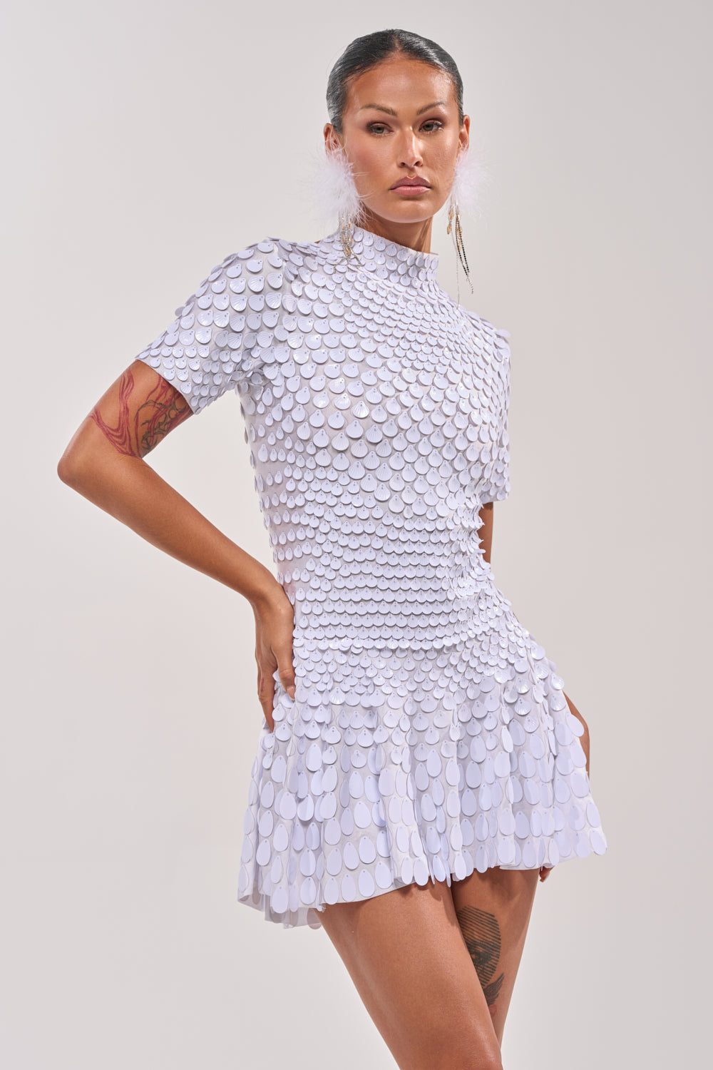 A woman poses with one hand on her hip in the FAR FROM BASIC SEQUIN MINI DRESS IN WHITE, a short-sleeved, high-neck fit-and-flare mini dress decorated with layered circular sequins, set against a plain light background. 1