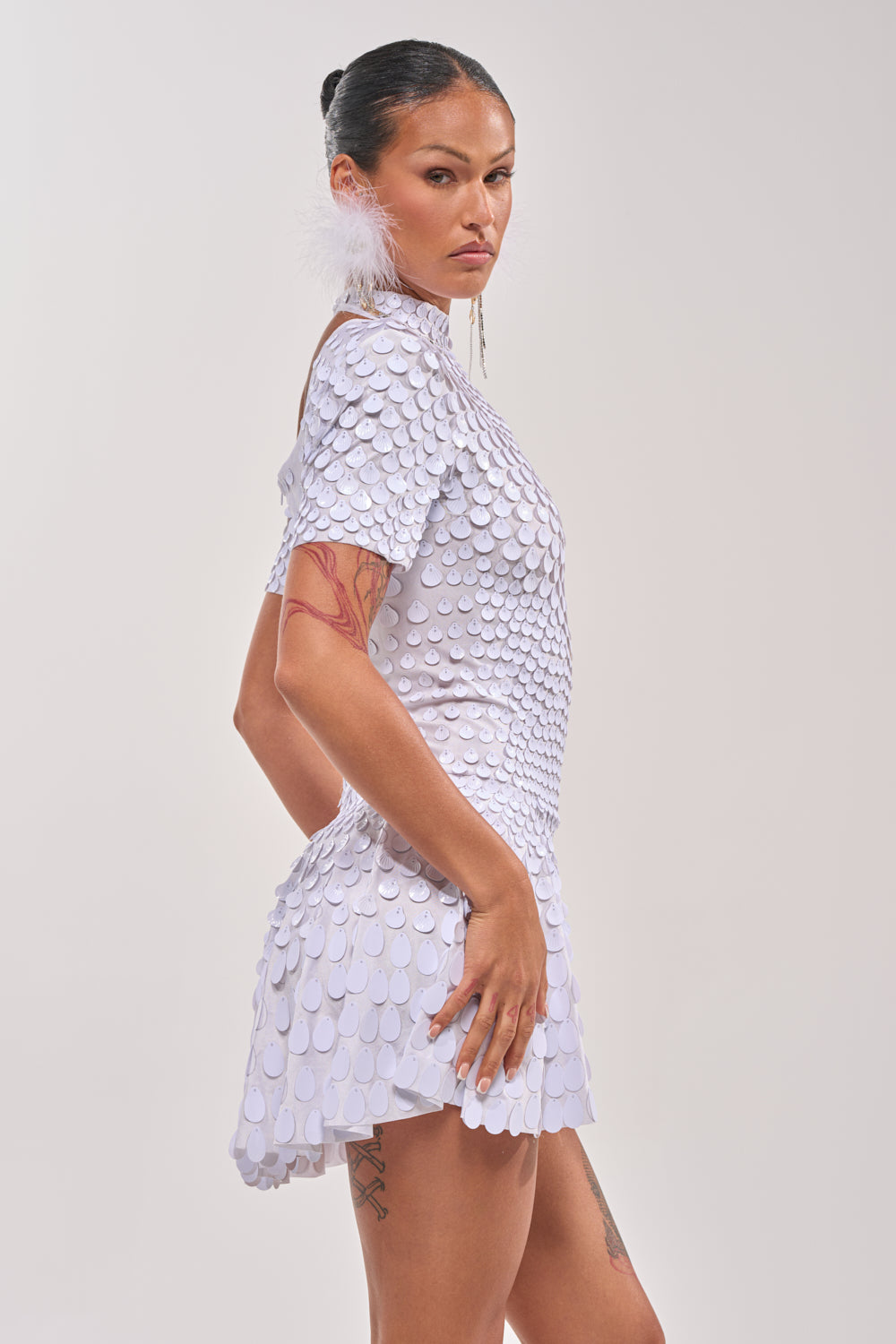 A person with long dark hair in a slicked-back bun wears the FAR FROM BASIC SEQUIN MINI DRESS IN WHITE and feather earrings, standing sideways and looking forward seriously. Tattoos are visible on their arm and leg. 3
