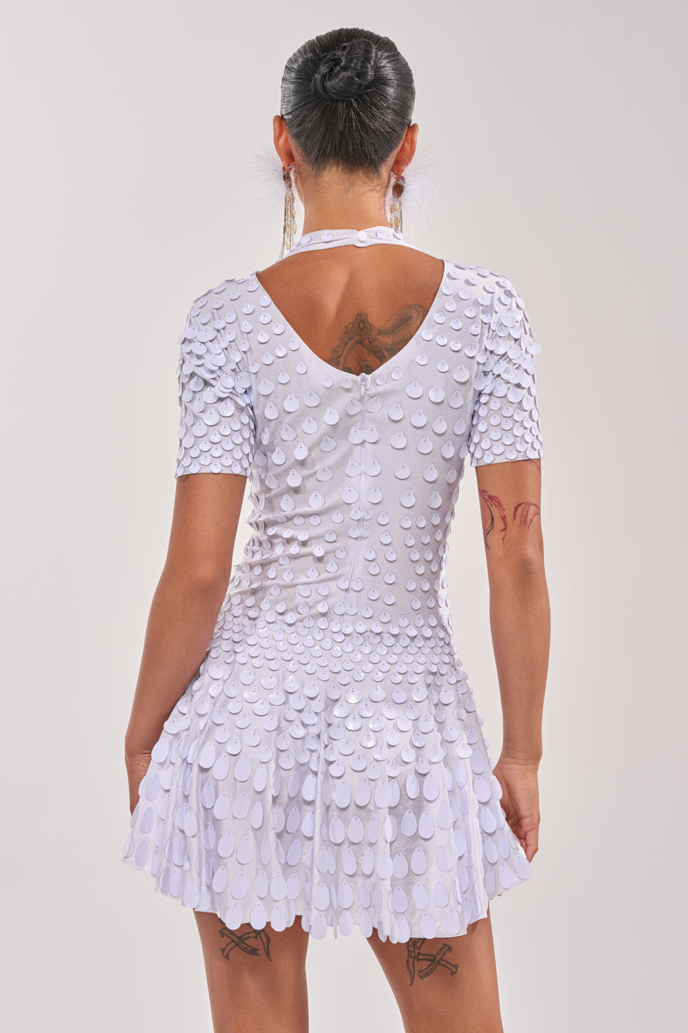 A person with dark hair in a bun, visible tattoos, and wearing the FAR FROM BASIC SEQUIN MINI DRESS IN WHITE stands facing away against a plain background. 4