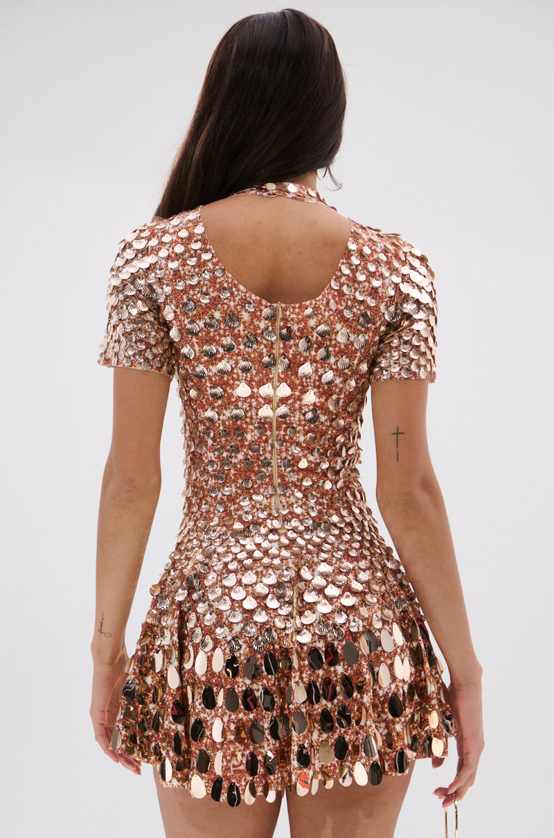 A woman with long dark hair faces away, wearing the FAR FROM BASIC SEQUIN MINI DRESS. The shiny mini dress features shell embellishments, round metallic discs, a flared skater skirt, and a visible back zipper against a plain background.