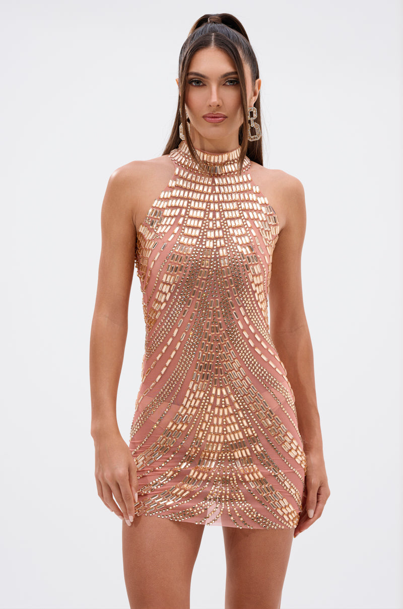 A woman with long, straight brown hair in a high half-ponytail wears the SHINE BRIGHT RHINESTONE MESH MINI DRESS IN ROSE GOLD—a sleeveless, fitted mini dress with gold geometric sequins on a nude base—standing against a plain white background.
