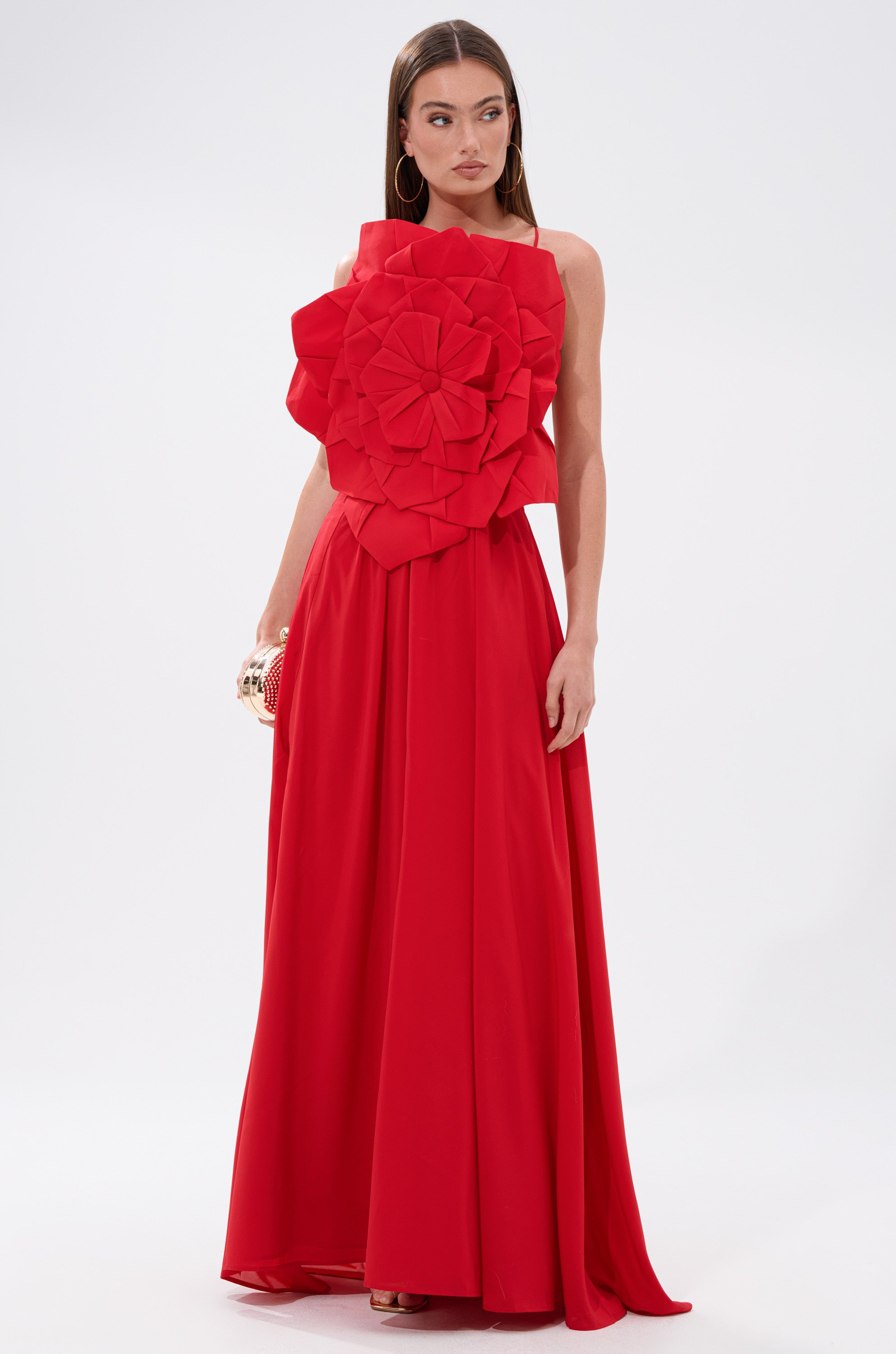 A woman models the LANIE MAXI DRESS WITH FLOWER APPLIQUE, a bold red gown featuring a sculpted flower detail on the bodice, paired with a small clutch against a plain white backdrop.