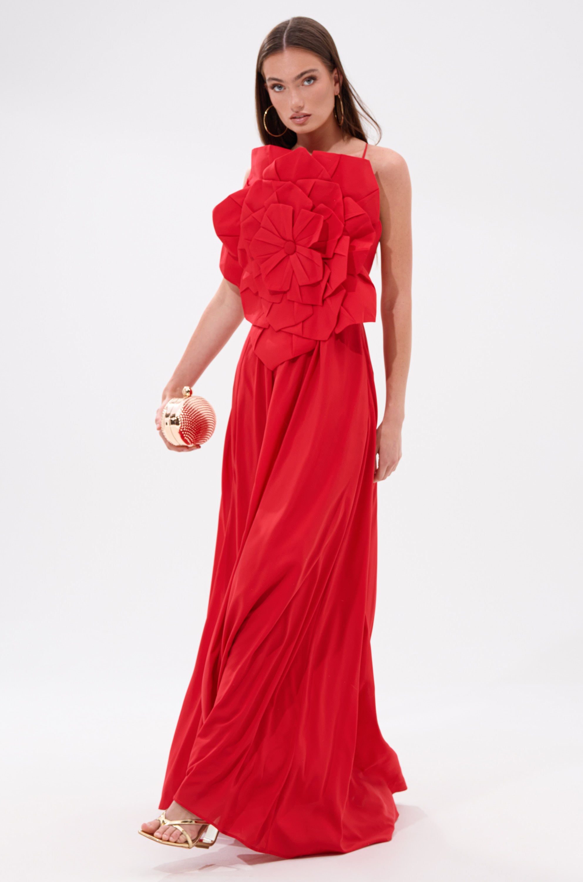 A woman models the LANIE MAXI DRESS WITH FLOWER APPLIQUE—a striking red dress with a sculpted flower on the bodice—paired with gold sandals and a small gold clutch, posing against a plain white background.