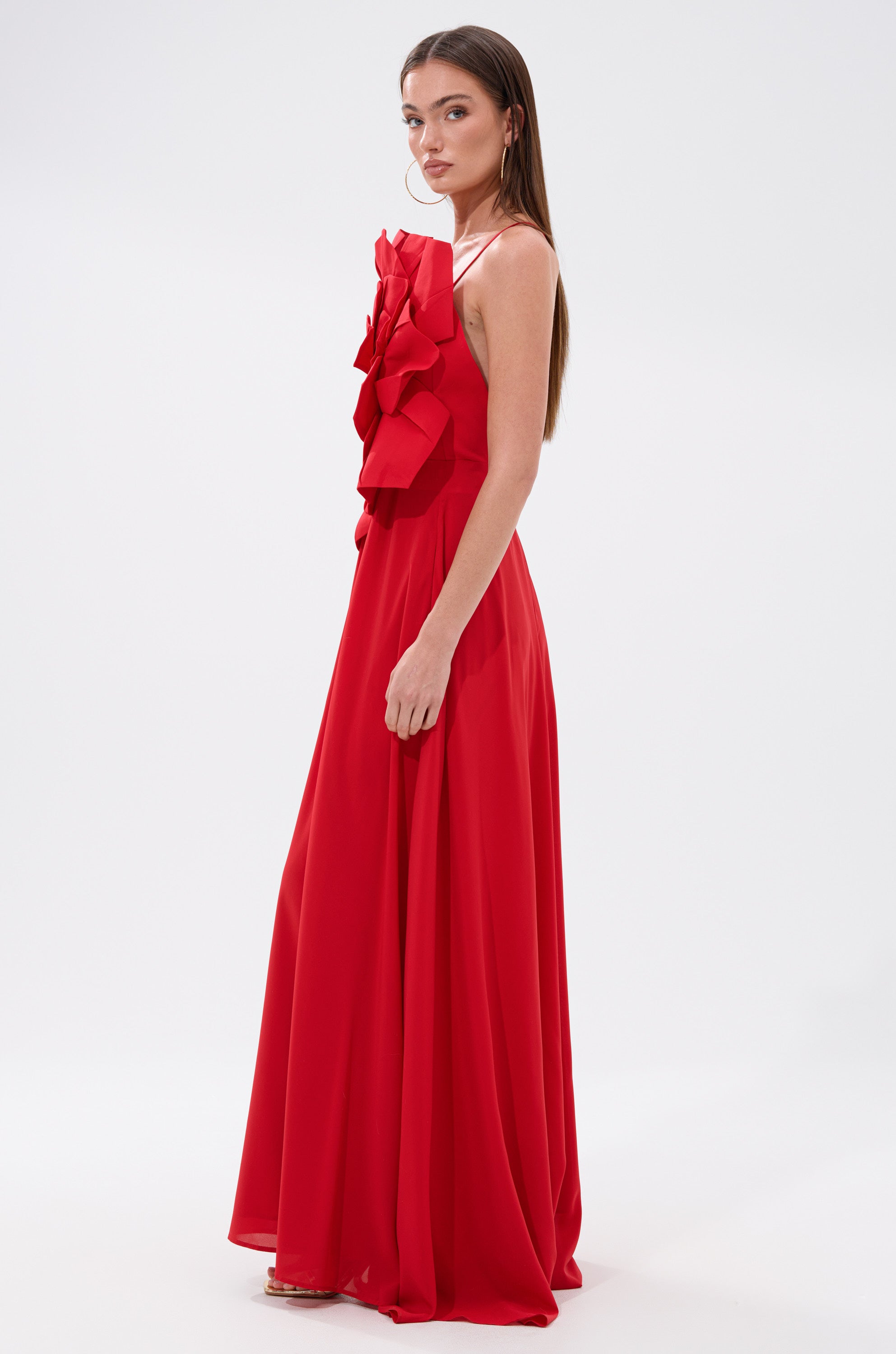 A woman in profile wears the LANIE MAXI DRESS WITH FLOWER APPLIQUE, a striking red maxi dress featuring front ruffle details. She has straight brown hair and neutral makeup, set against a plain white background.