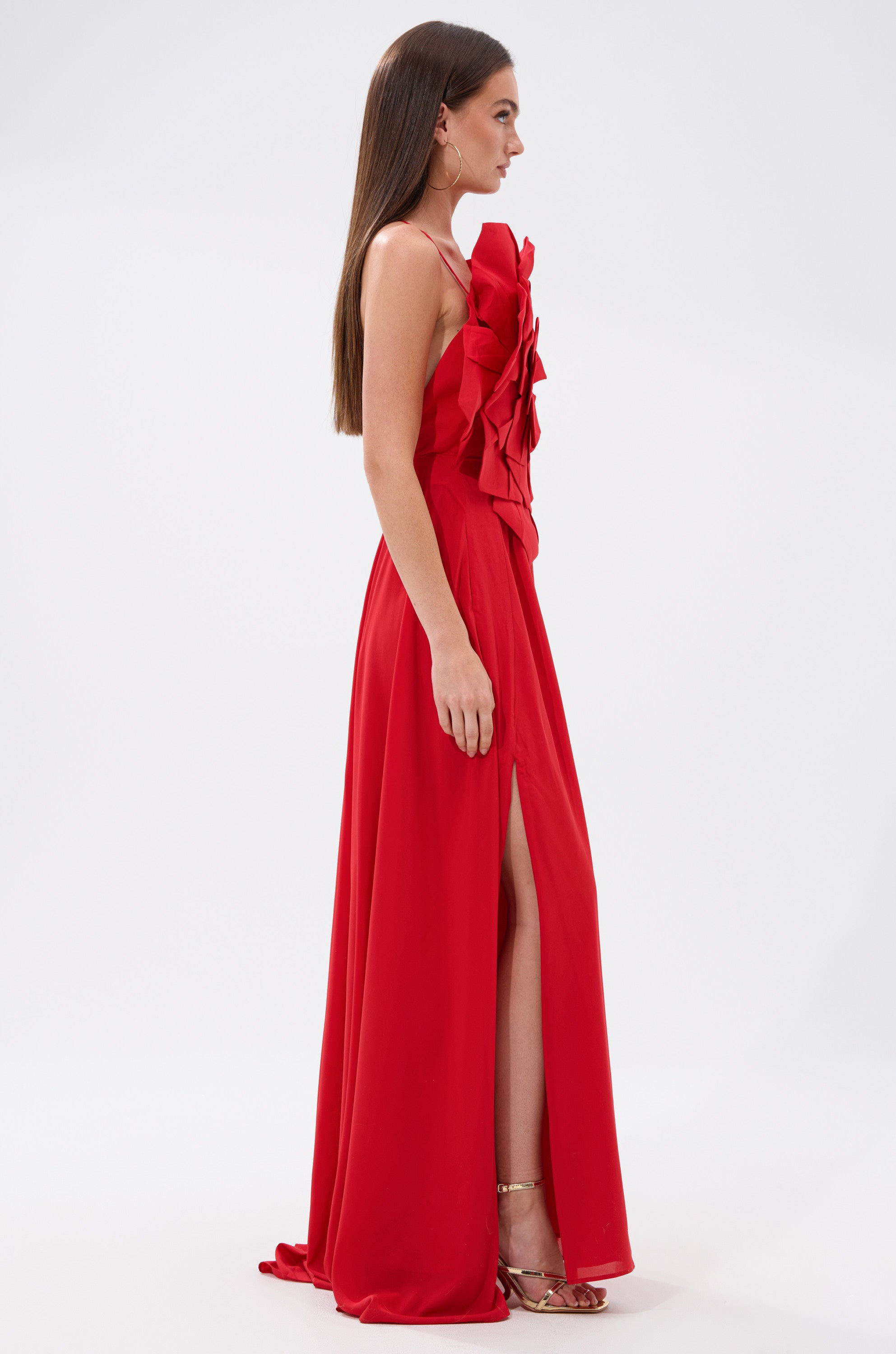 A woman stands in profile wearing the LANIE MAXI DRESS WITH FLOWER APPLIQUE, a sleeveless, floor-length red dress with ruffle bodice details and a high slit. She has straight brown hair and gold high-heeled sandals.