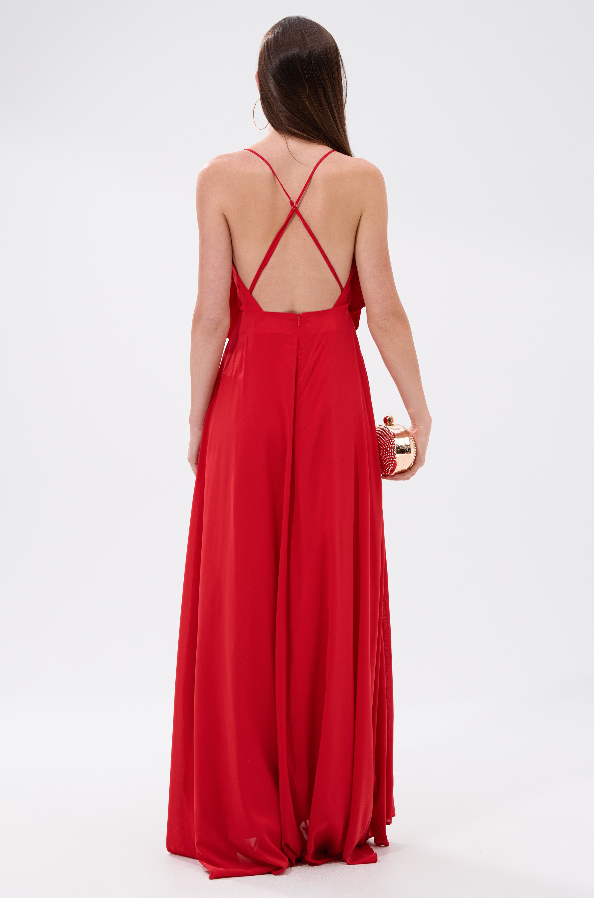 A woman with long brown hair wears the LANIE MAXI DRESS WITH FLOWER APPLIQUE, a red maxi dress with thin crisscross straps and an open back, holding a gold and red clutch against a plain white background.