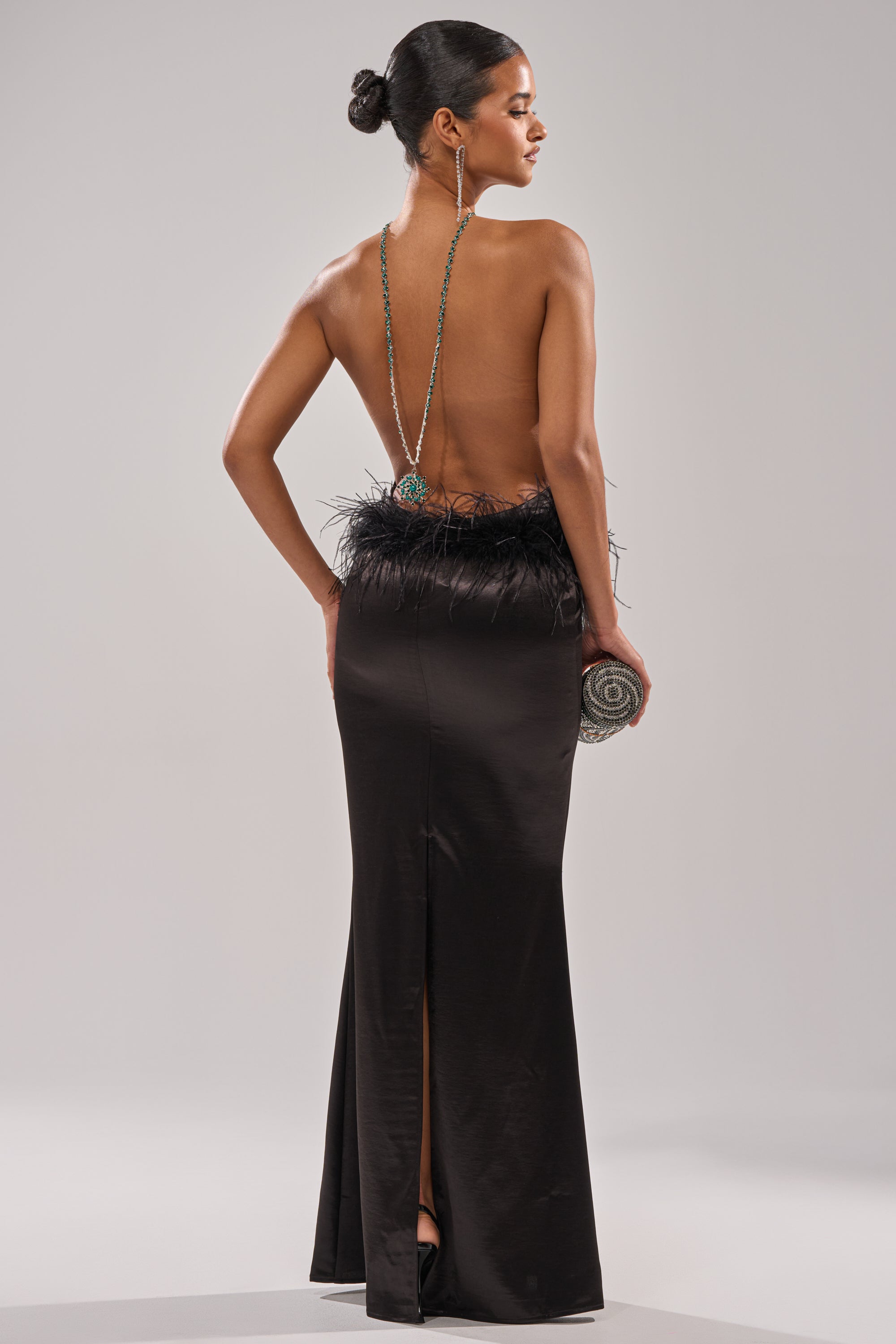 A woman wears the WHO IS SHE SATIN FEATHER MAXI DRESS IN BLACK, a floor-length, backless gown with feather accents at the waist, holding a silver round clutch and styling her hair in a neat bun as she faces away.