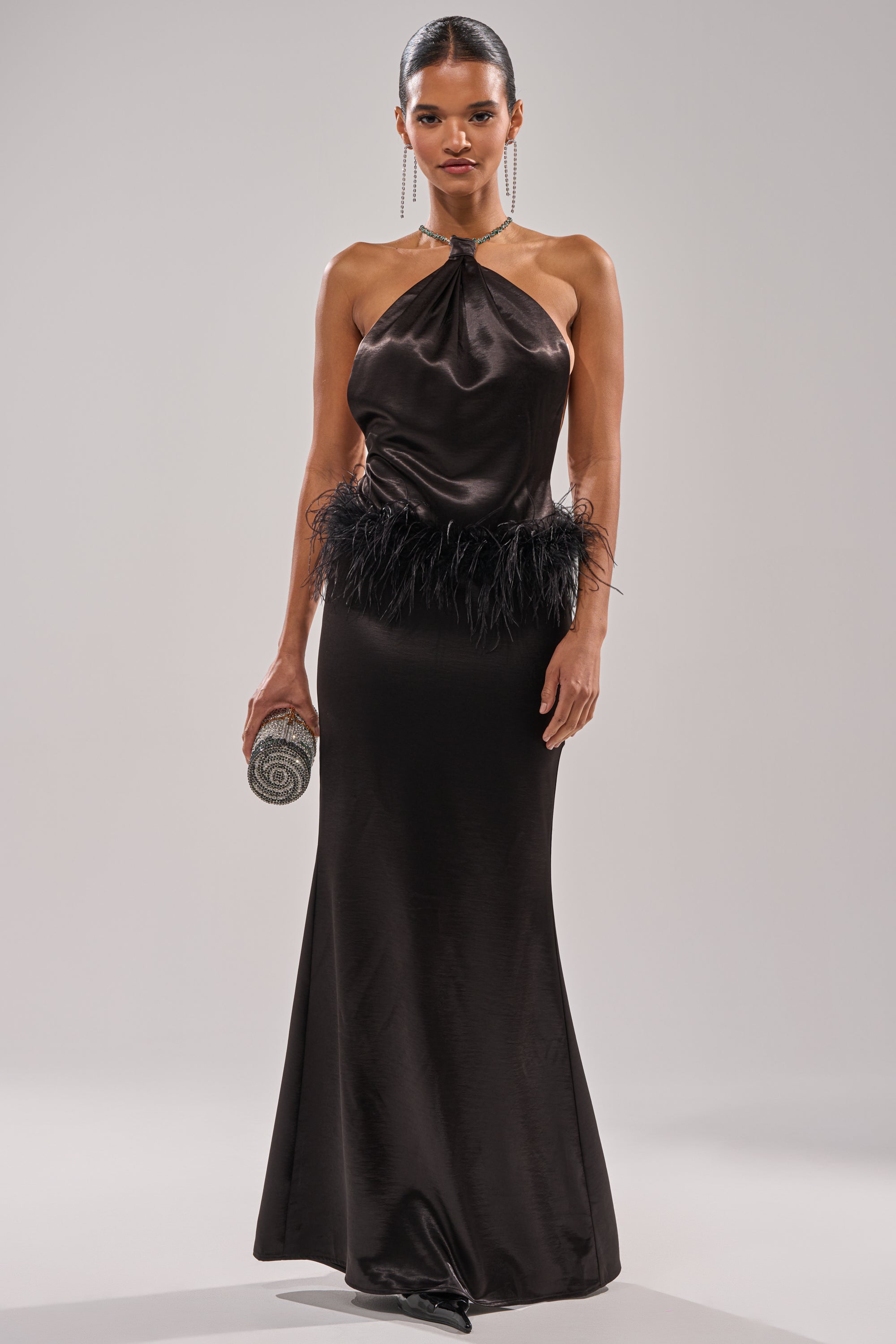 A woman wears the WHO IS SHE SATIN FEATHER MAXI DRESS IN BLACK, featuring a feather-accented waist, and holds a small round silver clutch while standing against a plain light gray background.