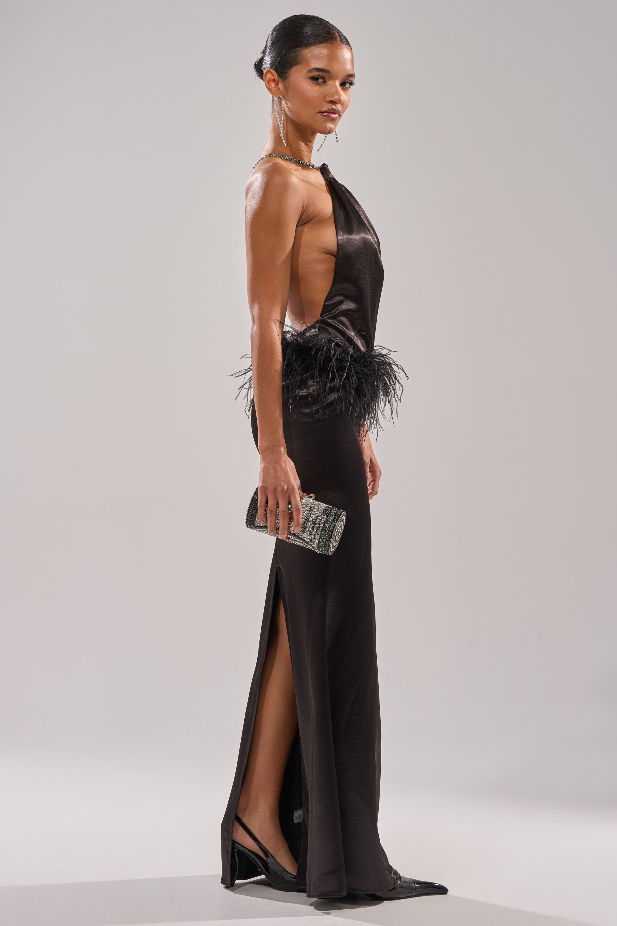 A woman confidently poses in the WHO IS SHE SATIN FEATHER MAXI DRESS IN BLACK, featuring a feather-accented waist. She completes her look with black heels, statement jewelry, and a small embellished clutch against a light background.