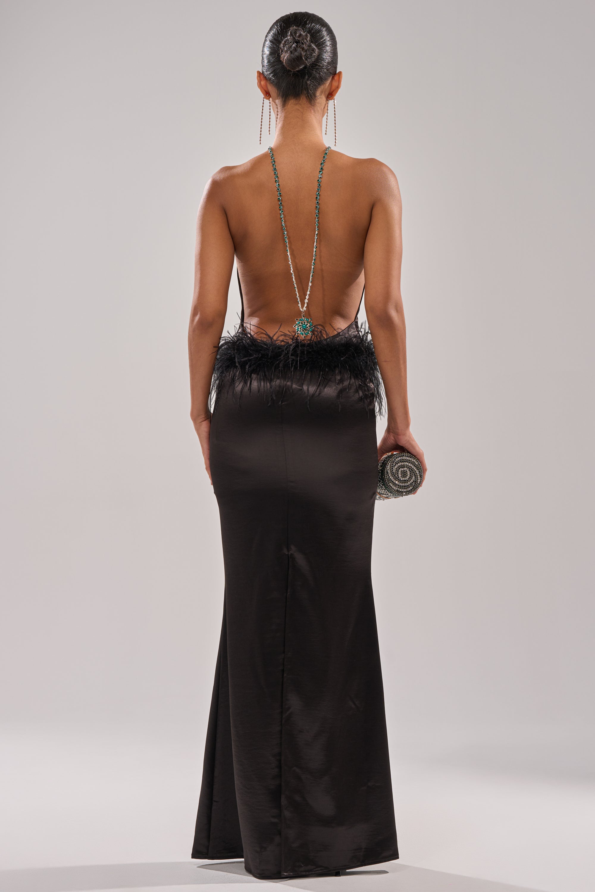 A woman with a sleek bun wears the WHO IS SHE SATIN FEATHER MAXI DRESS IN BLACK, featuring a dramatic backless cut, feather-trimmed waist, and holds a round clutch. A long necklace drapes elegantly down her back.