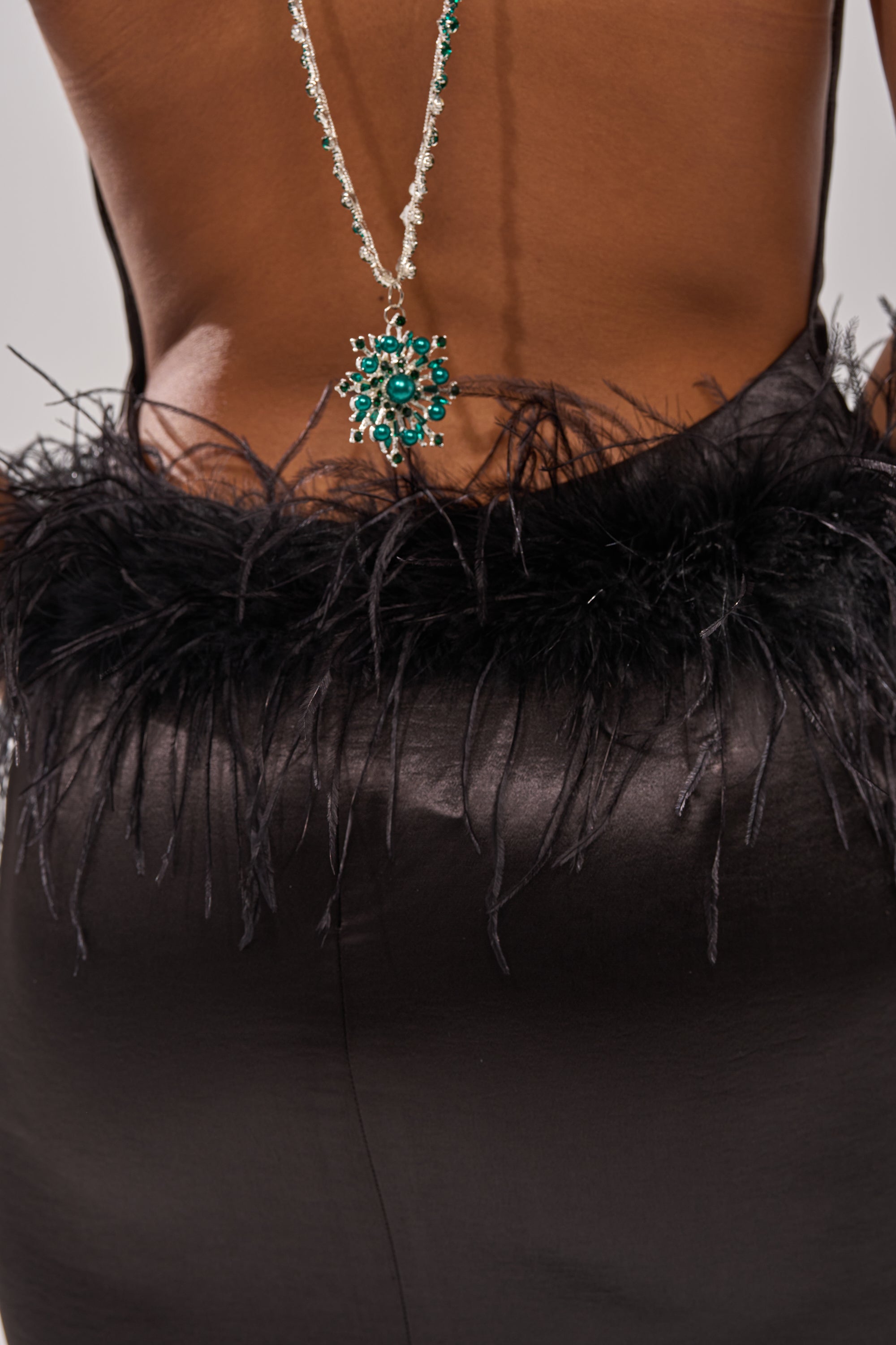 A close-up of a person’s lower back wearing the WHO IS SHE SATIN FEATHER MAXI DRESS IN BLACK, paired with an elegant necklace featuring green gemstones draped down their back.