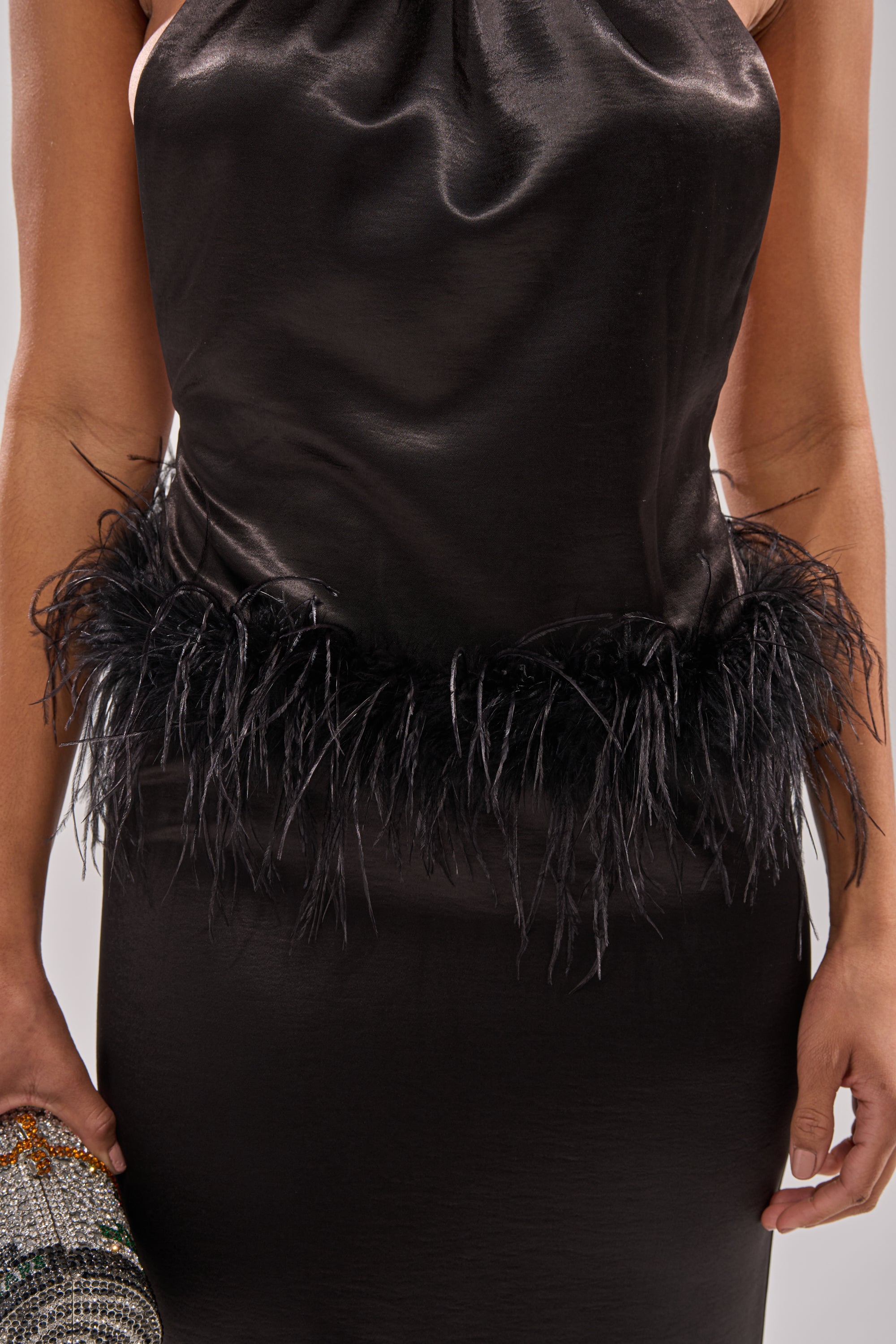 A person wearing the WHO IS SHE SATIN FEATHER MAXI DRESS IN BLACK, a sleeveless satin party dress with feather waist trim, holds a sparkly clutch. Only their torso and part of one arm are visible.
