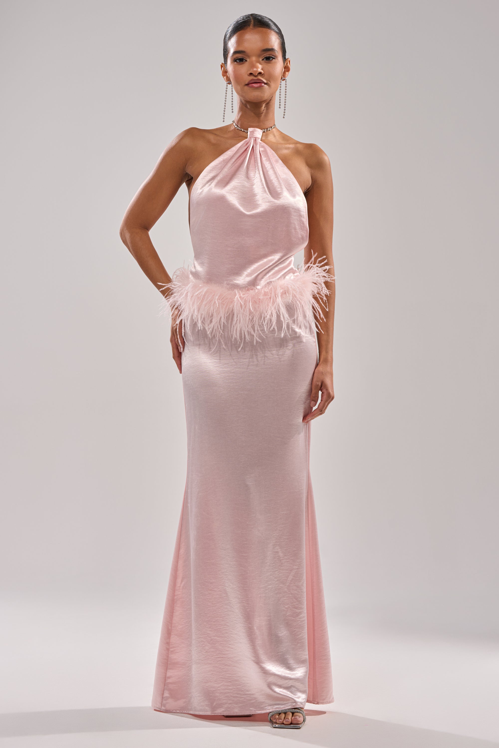 A woman poses gracefully in the WHO IS SHE SATIN FEATHER MAXI DRESS, a light pink gown with a gathered neckline and feathered waist trim, perfect for special events, against a plain light gray background.