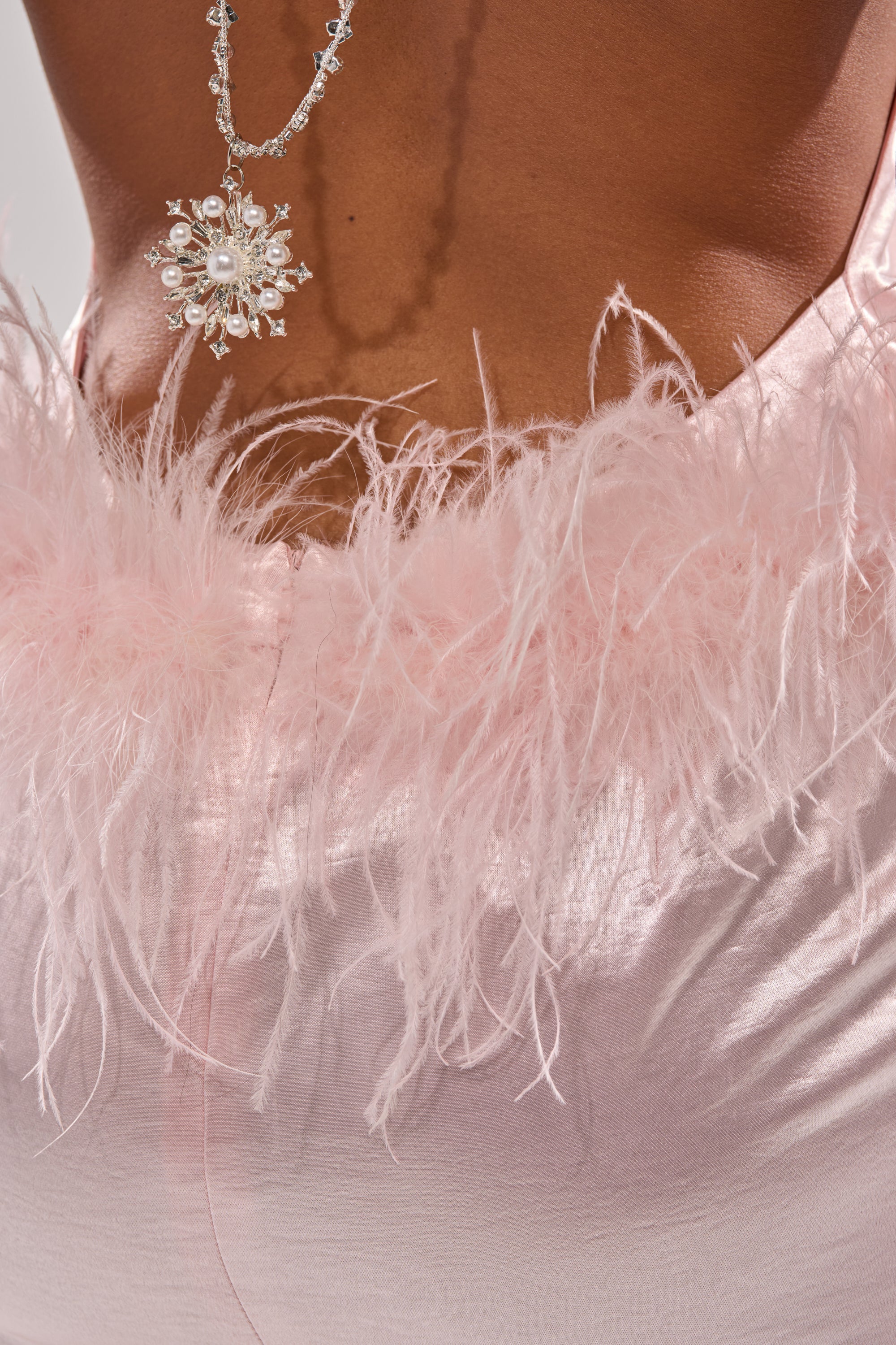 A close-up of someone wearing the WHO IS SHE SATIN FEATHER MAXI DRESS, featuring a shimmery pink satin fabric with pink feather trim. A silver snowflake necklace drapes down their open back—perfect for special occasions. 7