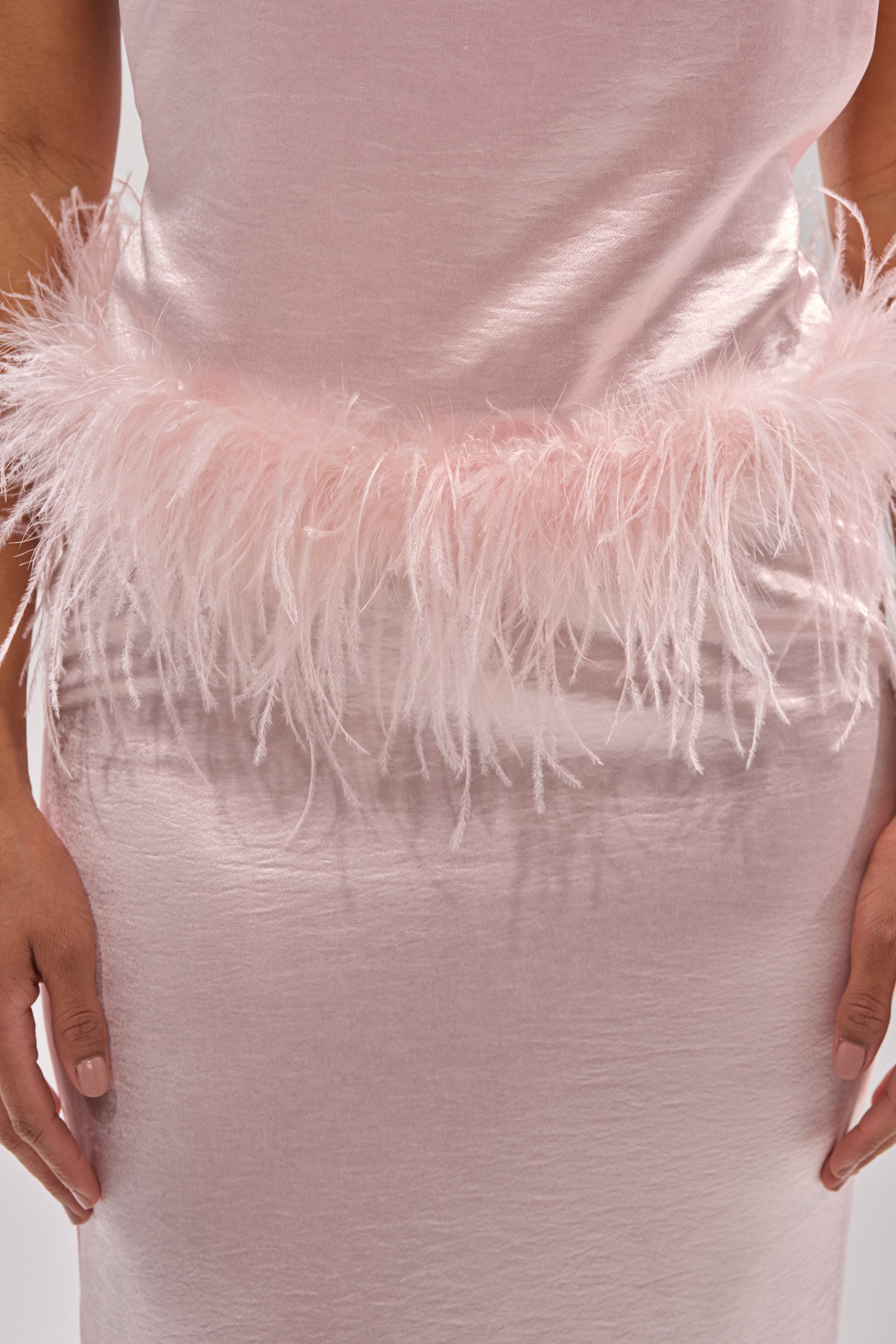 A close-up of someone wearing the WHO IS SHE SATIN FEATHER MAXI DRESS in pale pink, featuring a satin finish and feathered waist trim. Their hands rest at their sides, showcasing this elegant choice for special occasions. 8