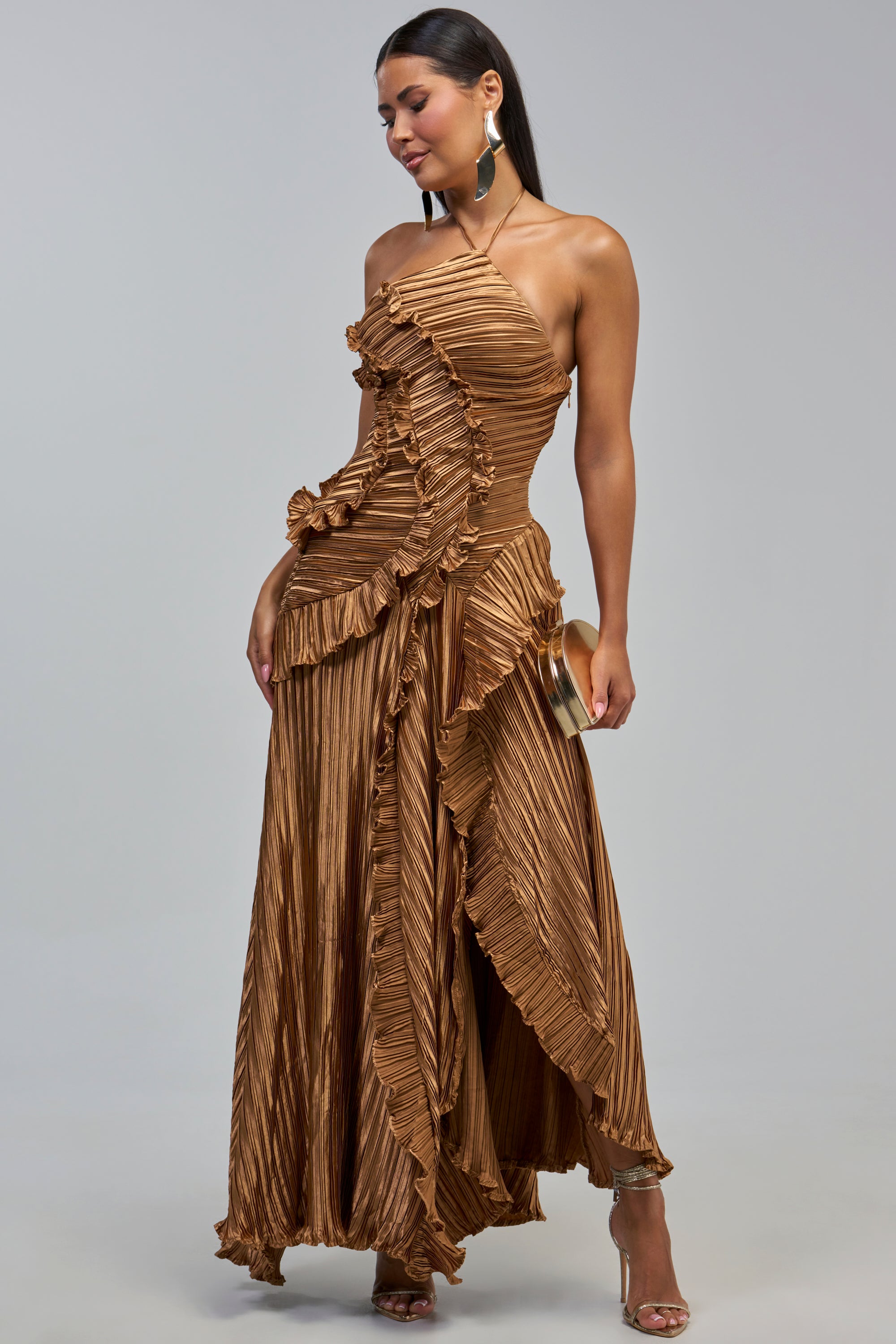A woman showcases the CELESTE RUFFLE MAXI DRESS IN BRONZE—a metallic bronze, pleated, asymmetrical ruffle one-shoulder formal dress—paired with a small clutch and high-heeled sandals against a plain background.
