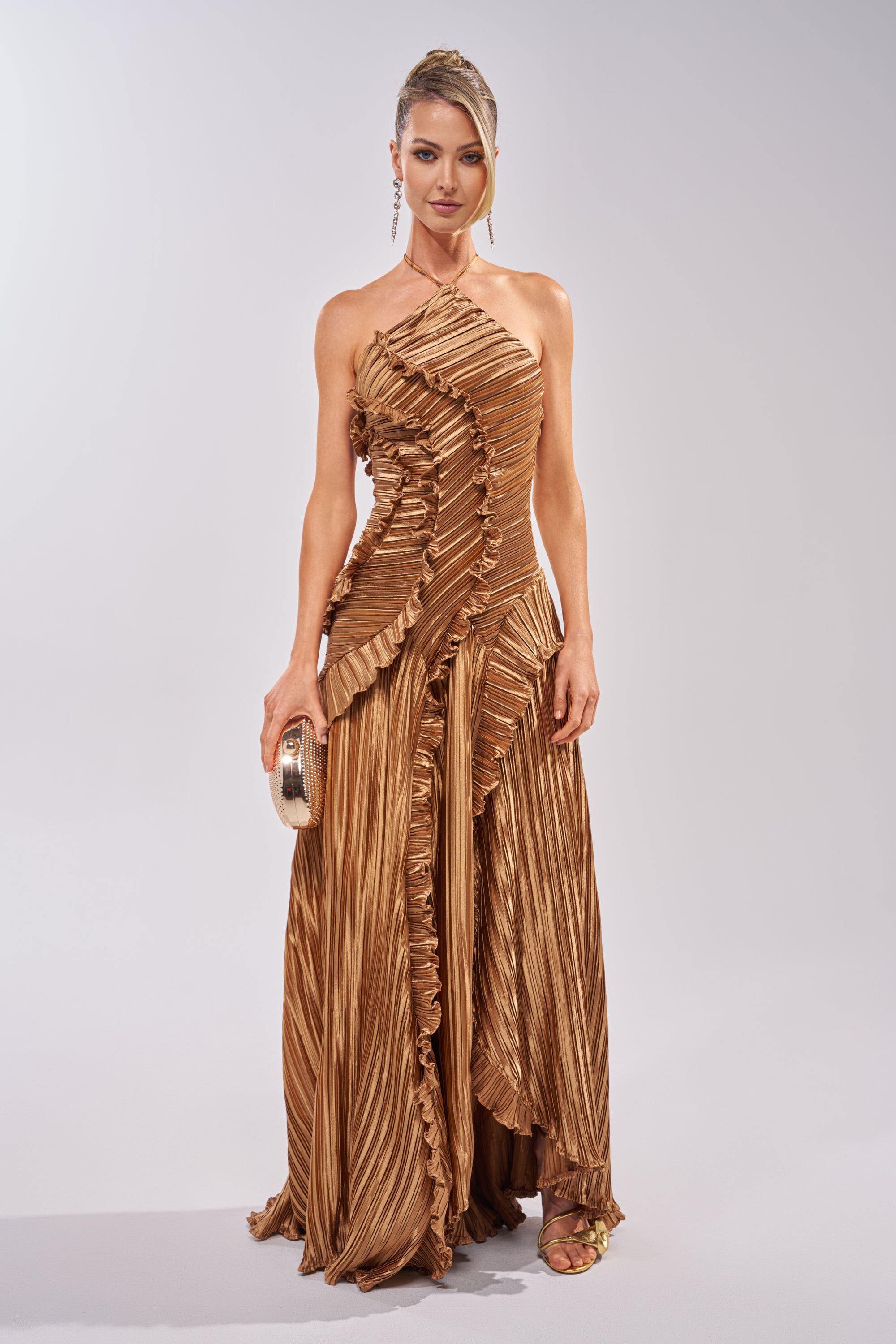 A woman stands against a plain background in the CELESTE RUFFLE MAXI DRESS IN BRONZE—a pleated, one-shoulder gown with sculptural ruffle detailing—paired with a gold clutch, earrings, strappy sandals, and an elegant updo. 1