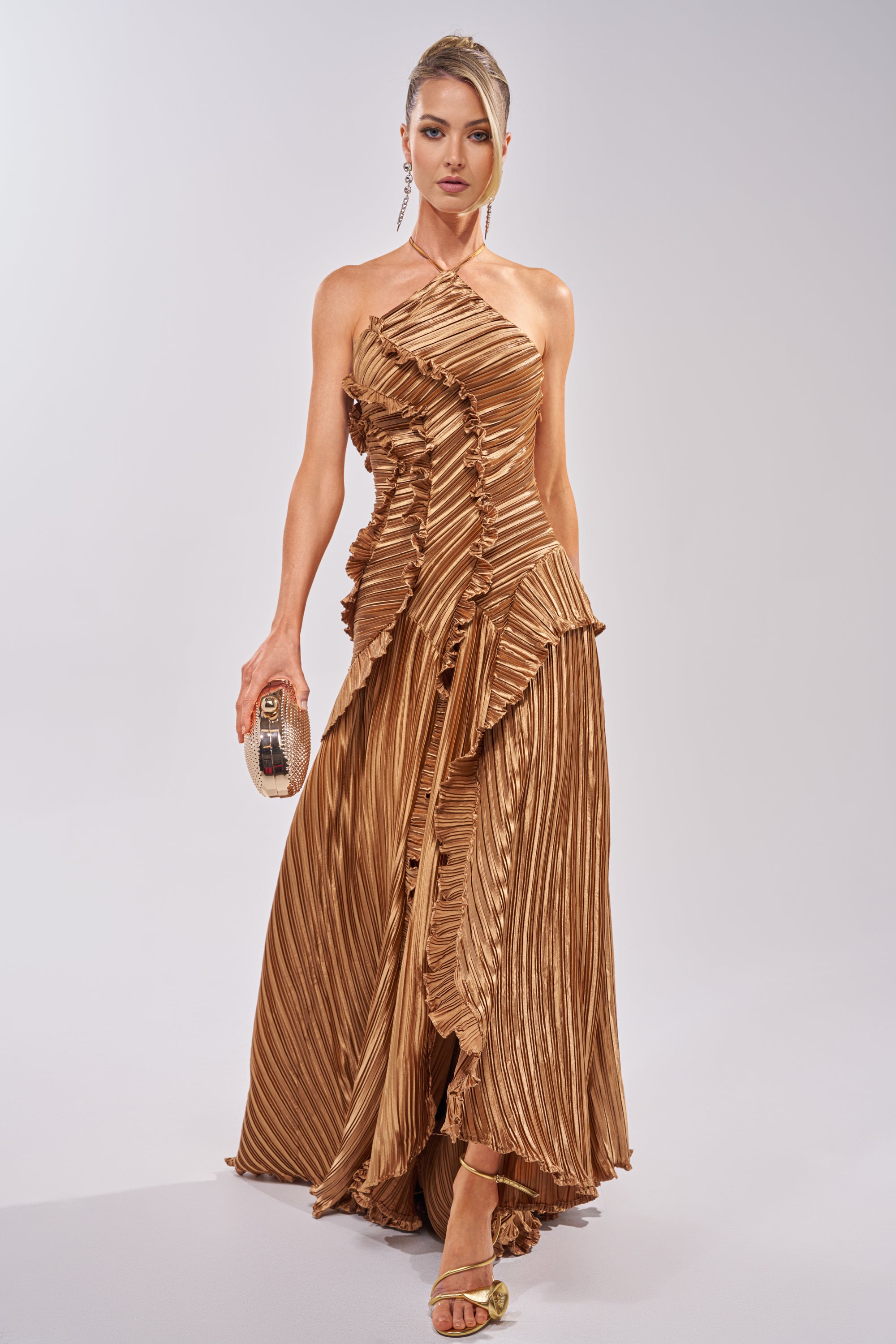 A woman in the CELESTE RUFFLE MAXI DRESS IN BRONZE, featuring pleats and sculptural ruffles, stands against a light background with a small clutch and strappy heels, her hair in an elegant updo. 2