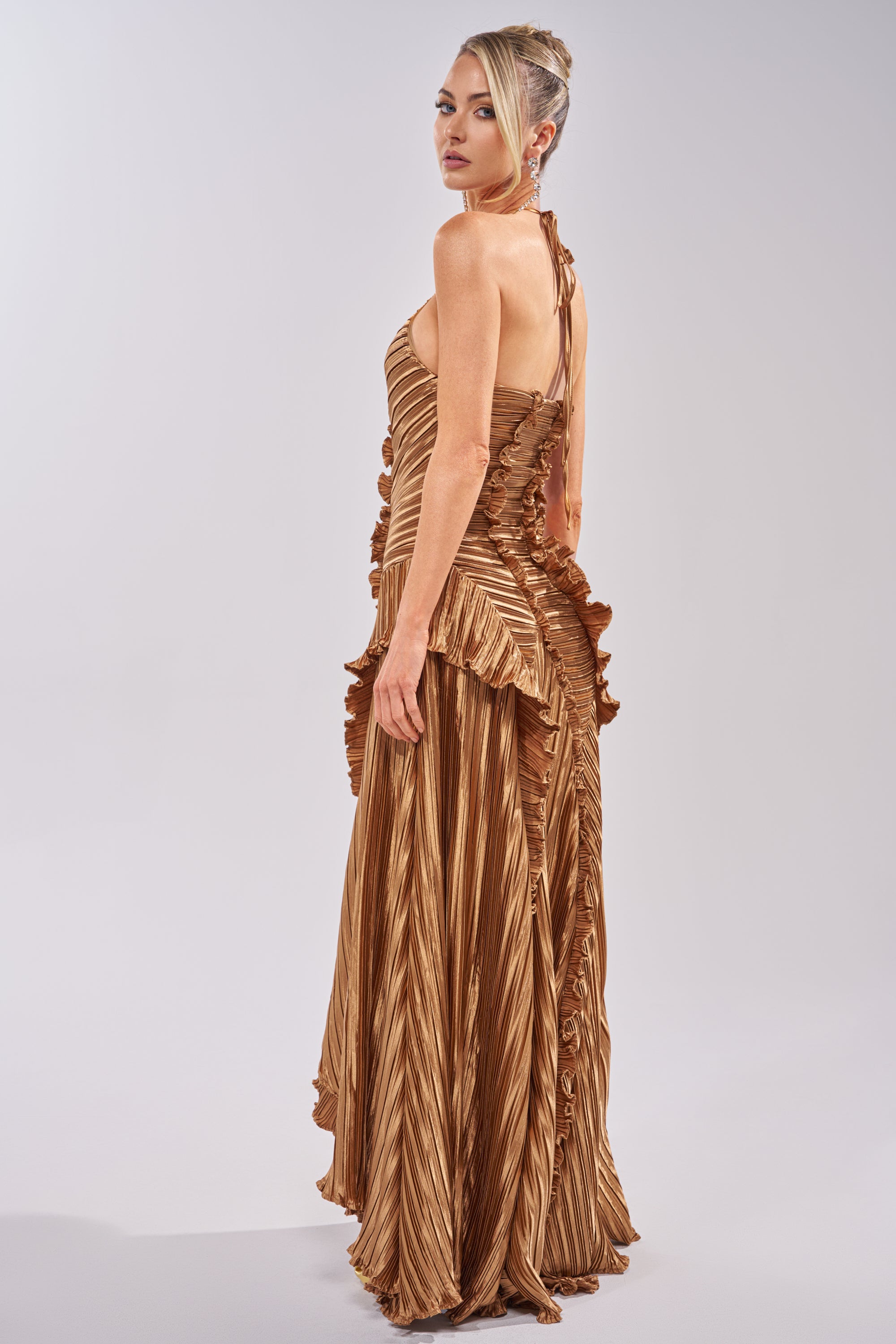 A woman in the CELESTE RUFFLE MAXI DRESS IN BRONZE, featuring metallic gold pleats and ruffle details, stands turned to the side with her blonde hair in an elegant updo against a plain light background. 4