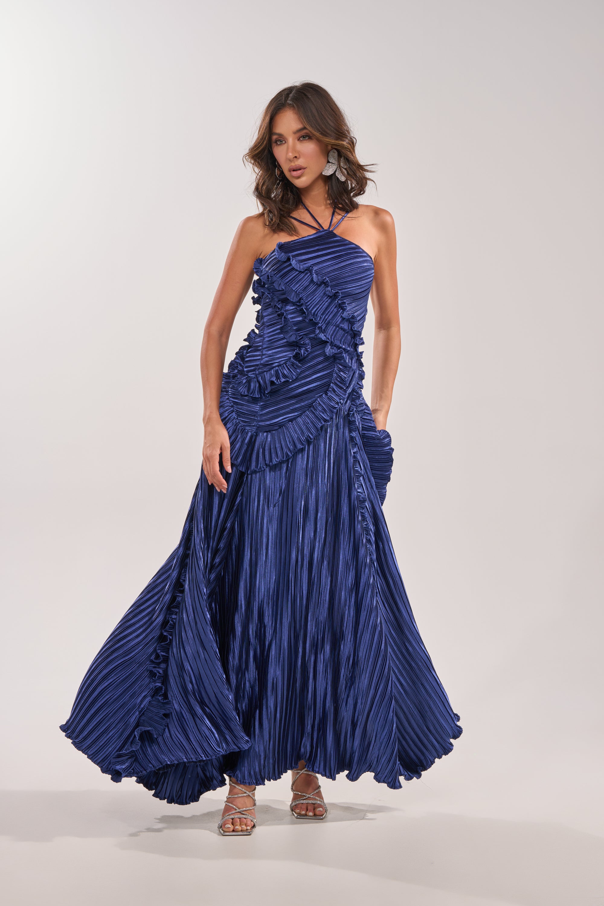 A woman models the CELESTE RUFFLE MIDI DRESS, featuring elegant pleats and ruffle accents. She pairs it with strappy heels, her wavy brown hair completing the sophisticated look against a simple light background. 5