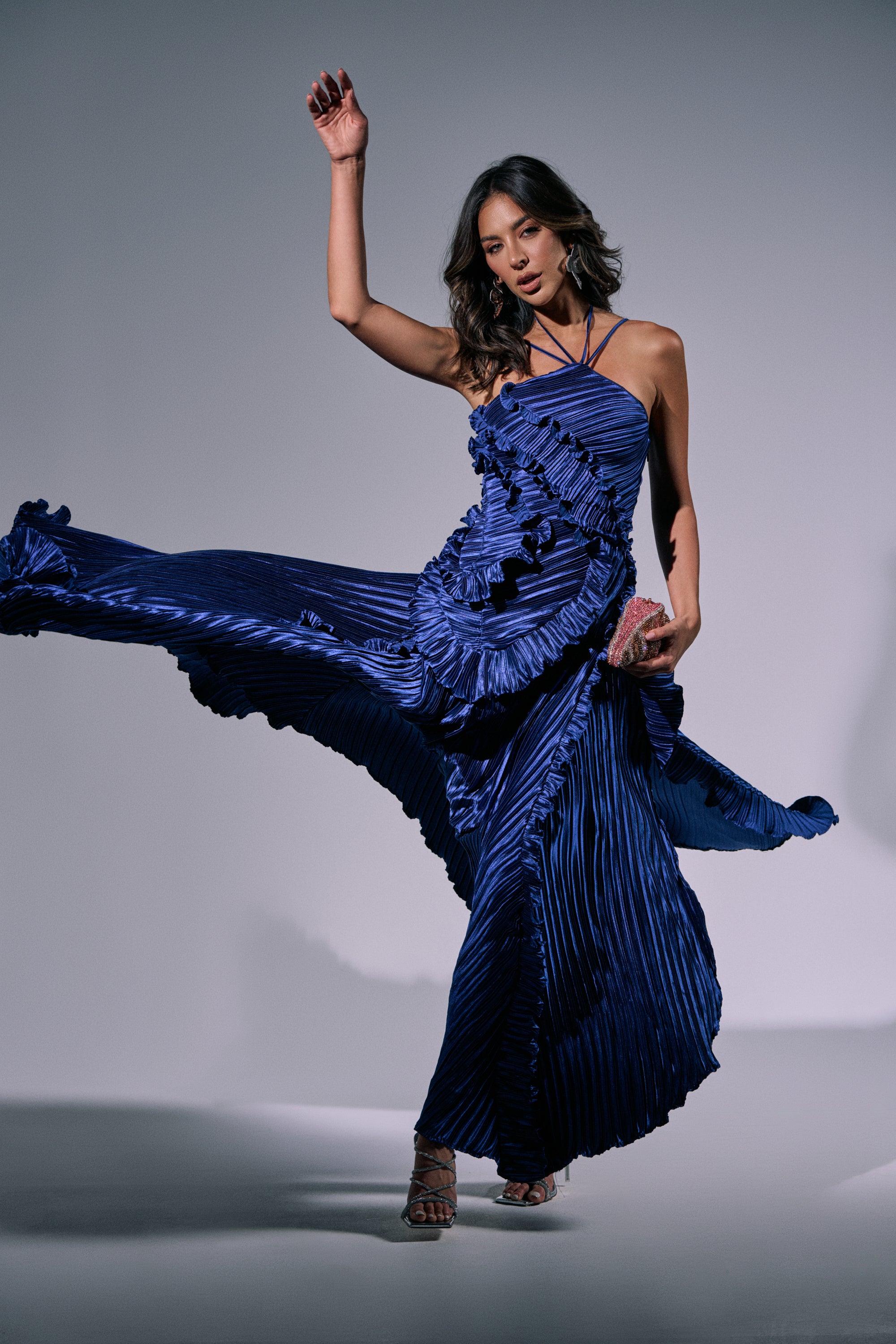 A woman poses dynamically in the CELESTE RUFFLE MIDI DRESS, lifting its hem to show off the ruffled details and halter neckline, creating movement against a plain, well-lit background. 4