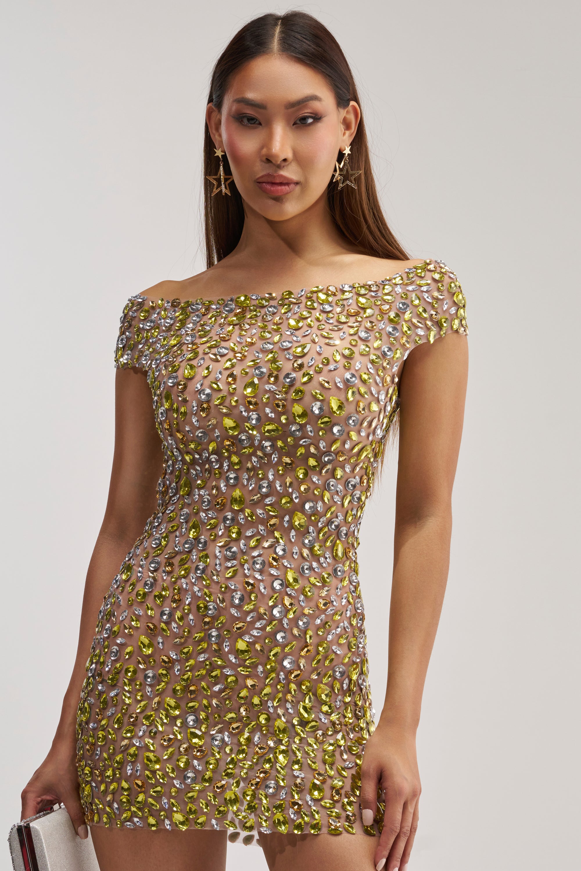 A woman in the HIGH STANDARDS RHINESTONE MINI DRESS—an off-the-shoulder, fitted mini dress adorned with yellow and silver gemstones—holds a clutch. She has long straight hair and accessorizes with star-shaped earrings.