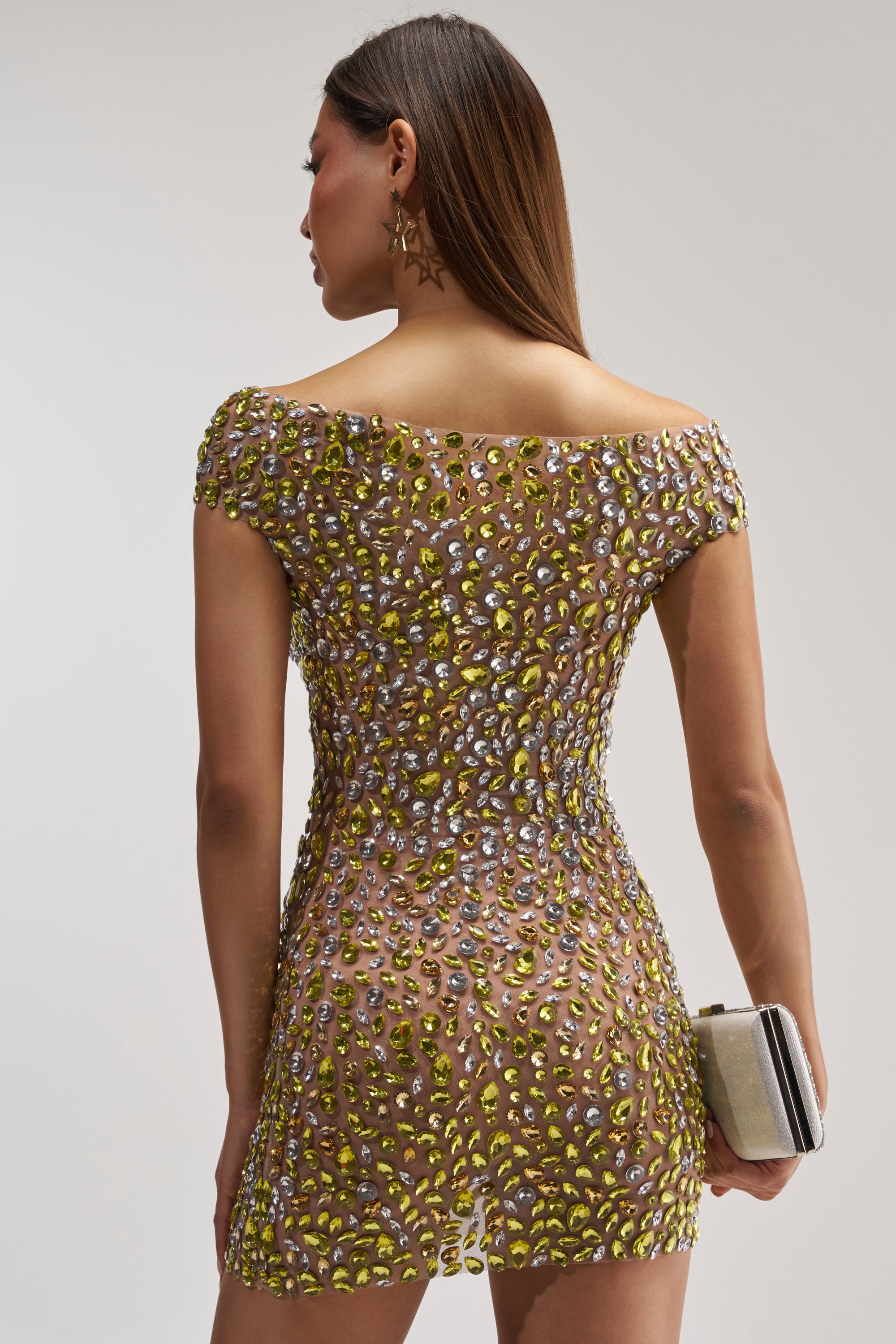Facing away, the woman wears the HIGH STANDARDS RHINESTONE MINI DRESS—a sheer gemstone mini adorned with yellow and silver rhinestone embellishments, making it an eye-catching birthday party choice. She holds a small clutch, her straight brown hair left loose and styled down, paired with star-shaped earrings against a neutral background.
