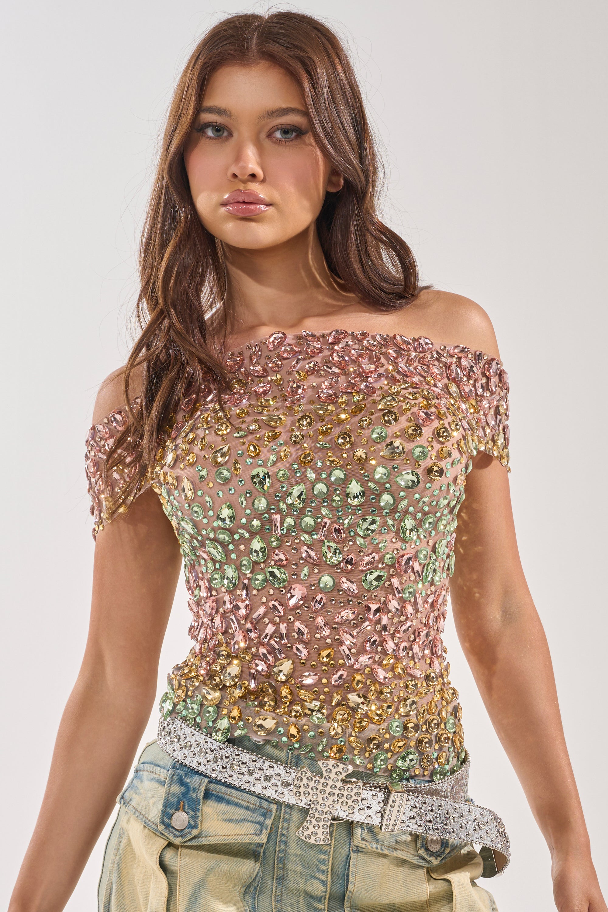 A woman with long brown hair wears the BLINGING OUT RHINESTONE TOP, an off-shoulder style with gold, green, and pink sequins—ideal for birthdays. She pairs it with light pants and a sparkly silver belt against a plain backdrop.