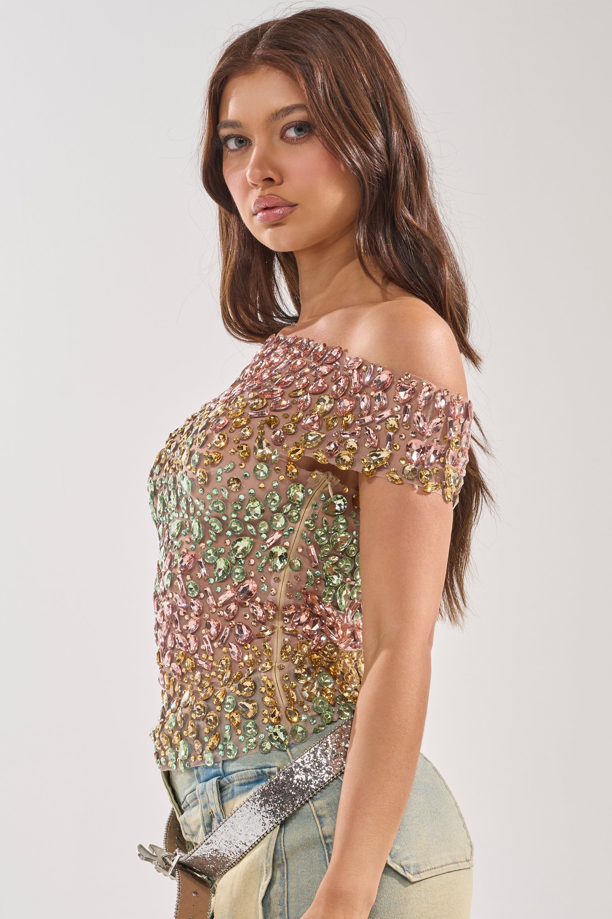 A woman with long brown hair wears the BLINGING OUT RHINESTONE TOP—an off-shoulder pastel piece adorned with colorful sequins and beads—plus light blue jeans and a glittery silver belt. She stands sideways, looking toward the camera.