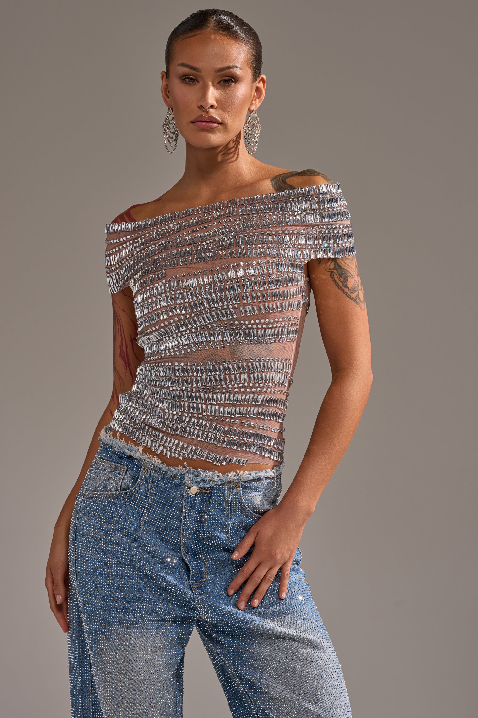 A woman stands confidently in the SHINE LIKE A DIAMOND RHINESTONE MESH BLOUSE with silver horizontal embellishments, paired with blue embellished jeans. She wears large dangling earrings, showcasing tattoos on her arms against a neutral background.
