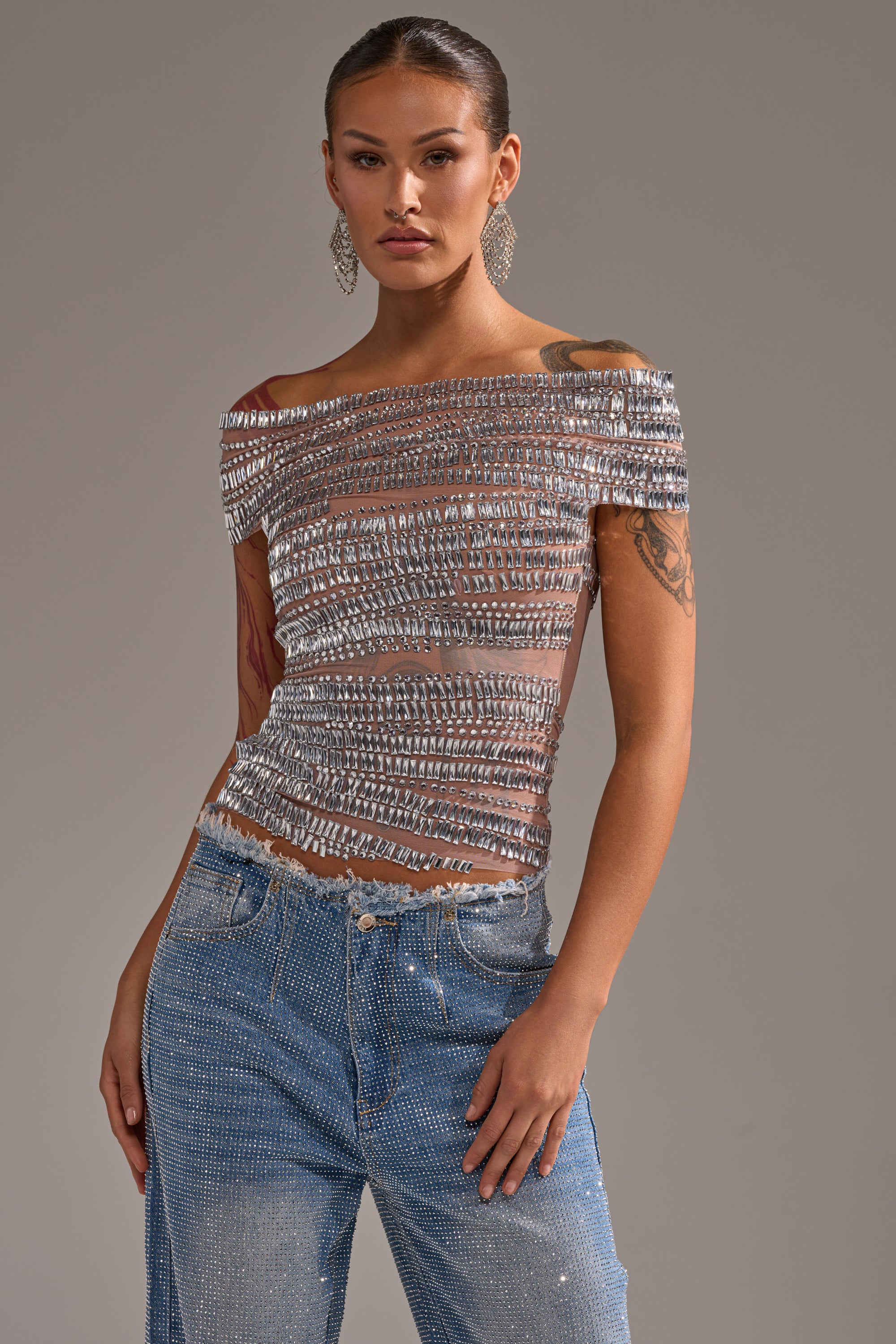A woman models the SHINE LIKE A DIAMOND RHINESTONE MESH BLOUSE, a short-sleeved, off-shoulder top with silver embellishments, paired with sparkly blue jeans and statement earrings for a chic, party-ready look against a plain background.