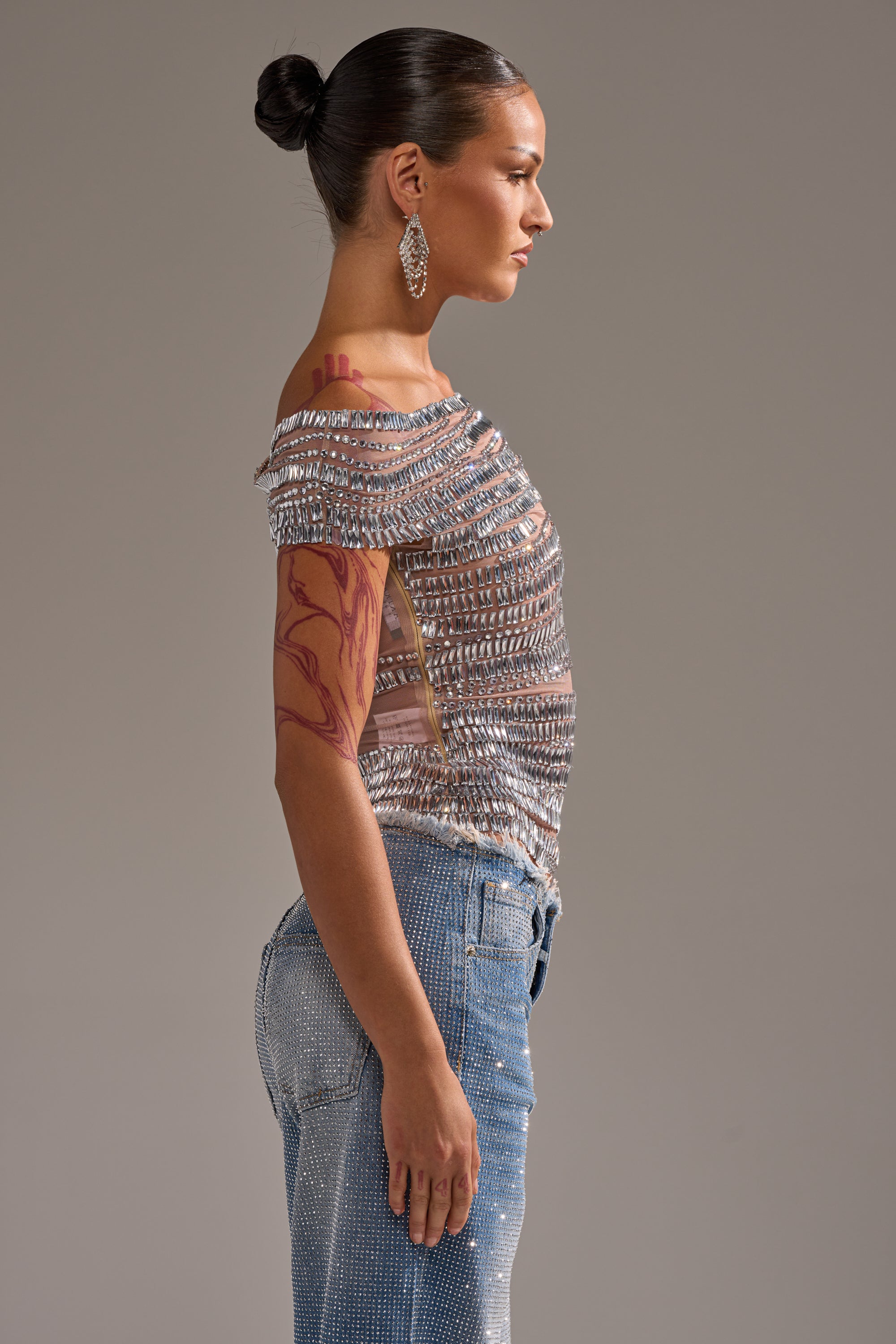 A woman in profile poses against a plain backdrop, wearing the SHINE LIKE A DIAMOND RHINESTONE MESH BLOUSE with sparkling jeans. Her hair is styled in a neat bun, accessorized with large earrings and a visible upper arm tattoo.