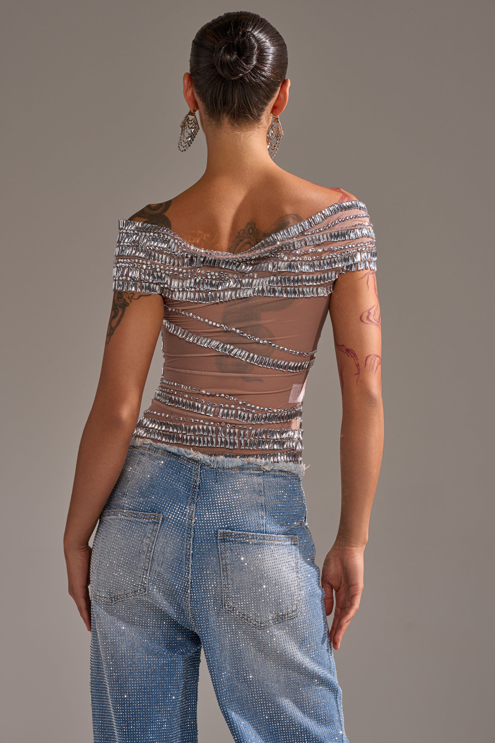 A person with their hair in a bun faces away, wearing the SHINE LIKE A DIAMOND RHINESTONE MESH BLOUSE and light blue rhinestone-studded jeans. They have visible tattoos and large earrings.