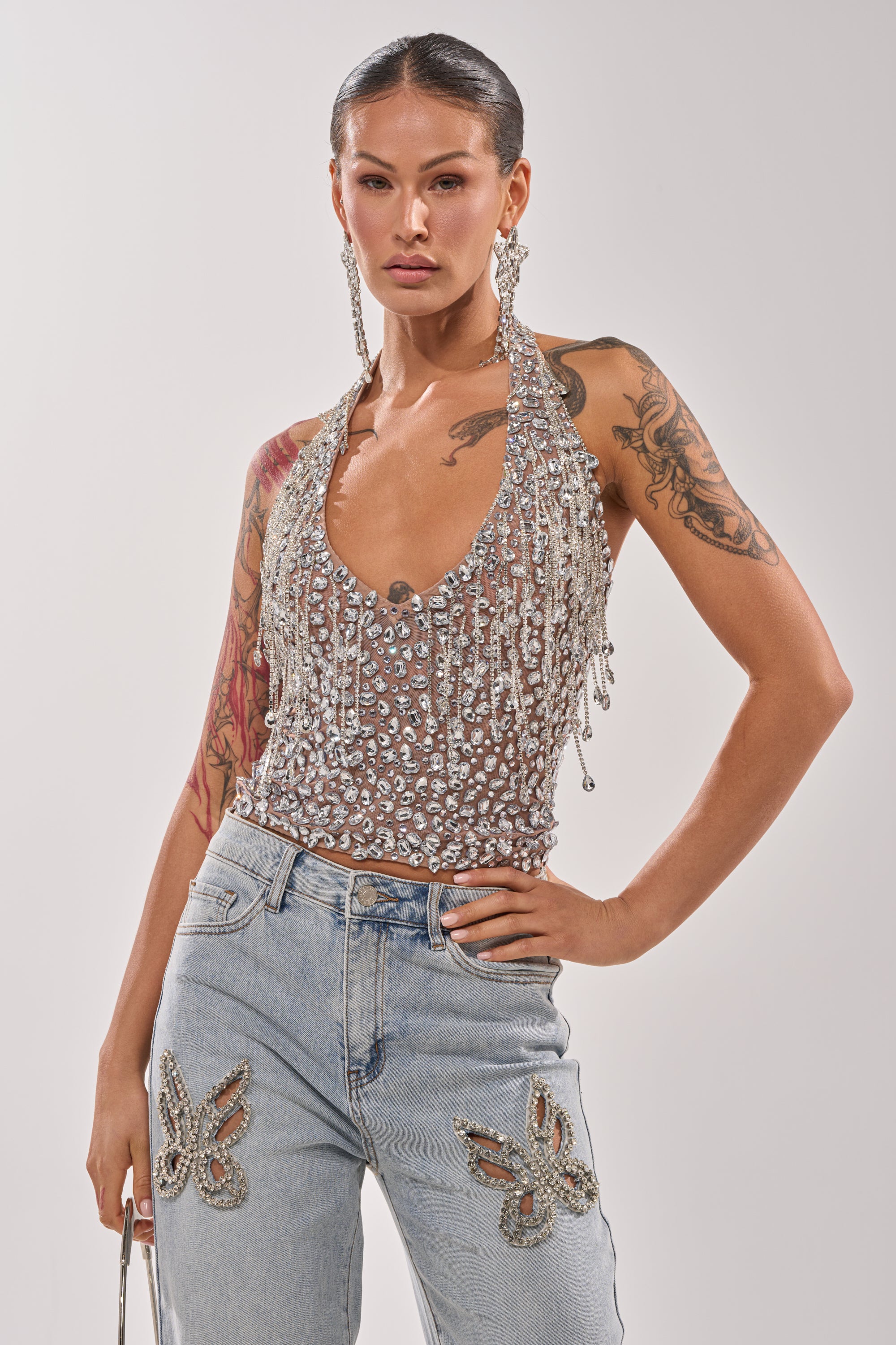 A person stands confidently in the HOTTEST ON THE BLOCK RHINESTONE BLOUSE with chain link fringe, large silver earrings, and butterfly-embellished light blue jeans, showing off their arm tattoos.