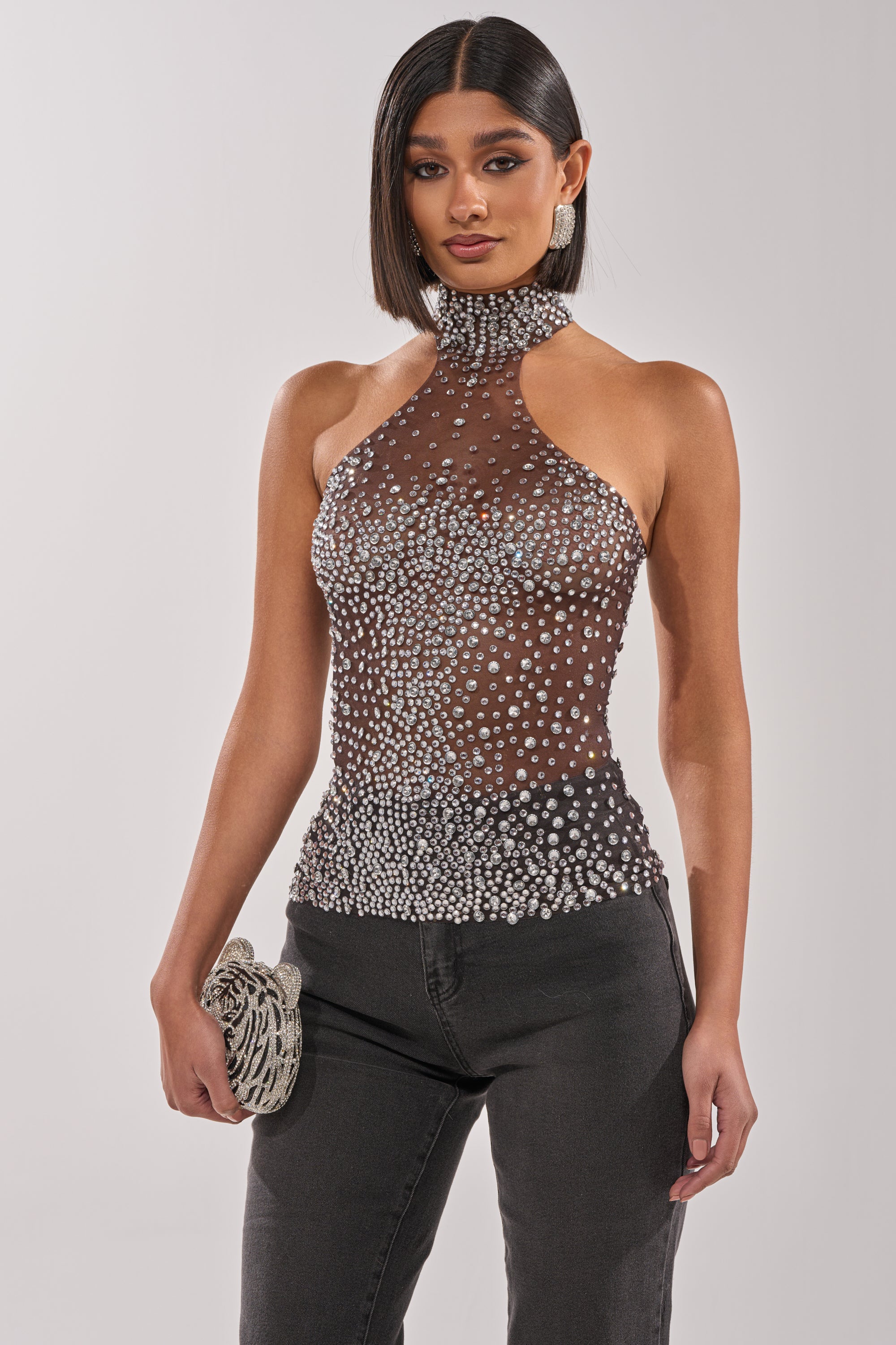 A woman with straight, shoulder-length hair wears the ALL YEAR ROUND SHEER RHINESTONE TOP, featuring sparkling rhinestones. She pairs it with black pants and carries a small silver clutch. 0