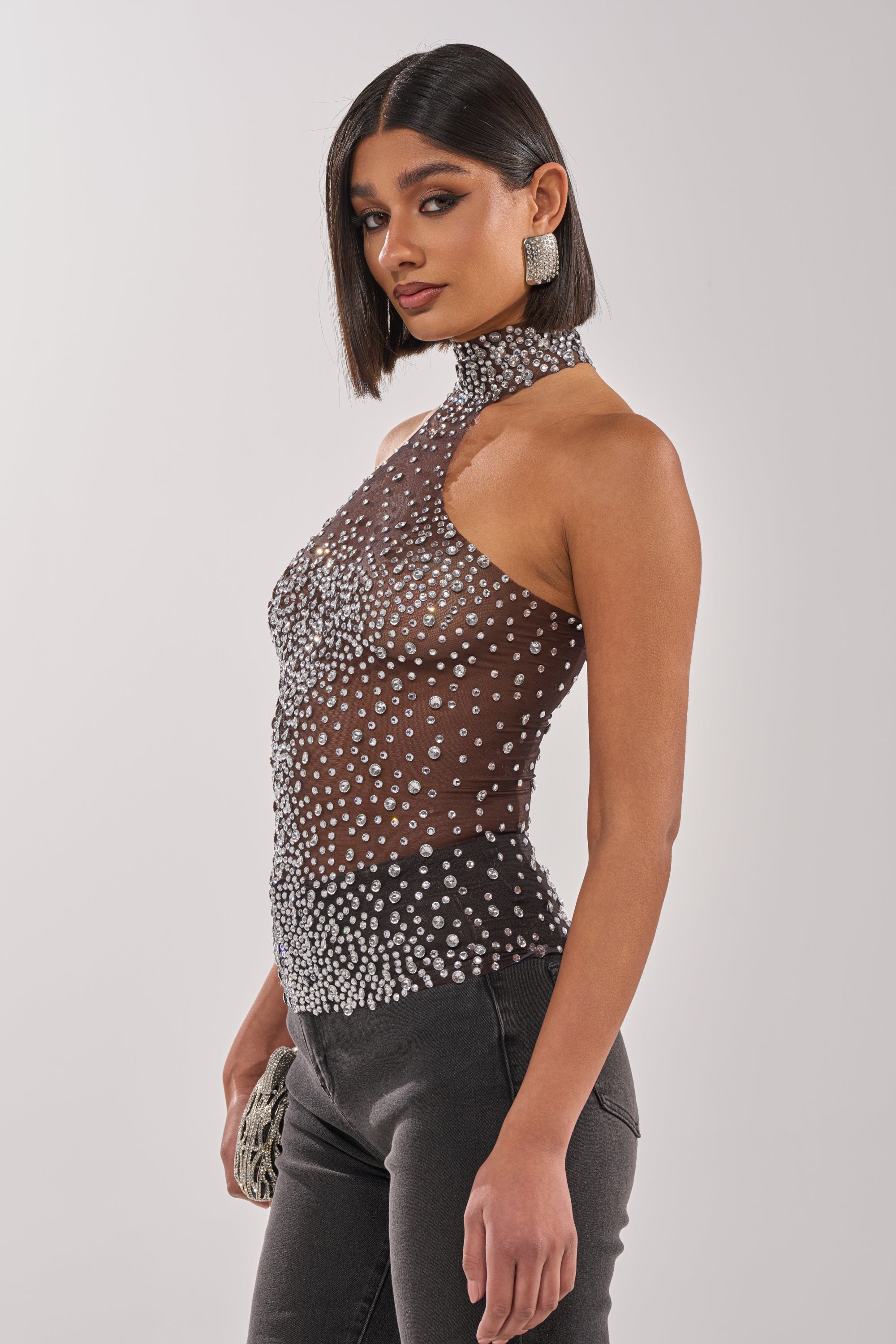 A woman with straight dark hair wears the ALL YEAR ROUND SHEER RHINESTONE TOP with black pants, holds a small clutch, and accessorizes with statement earrings while posing against a plain light background. 2