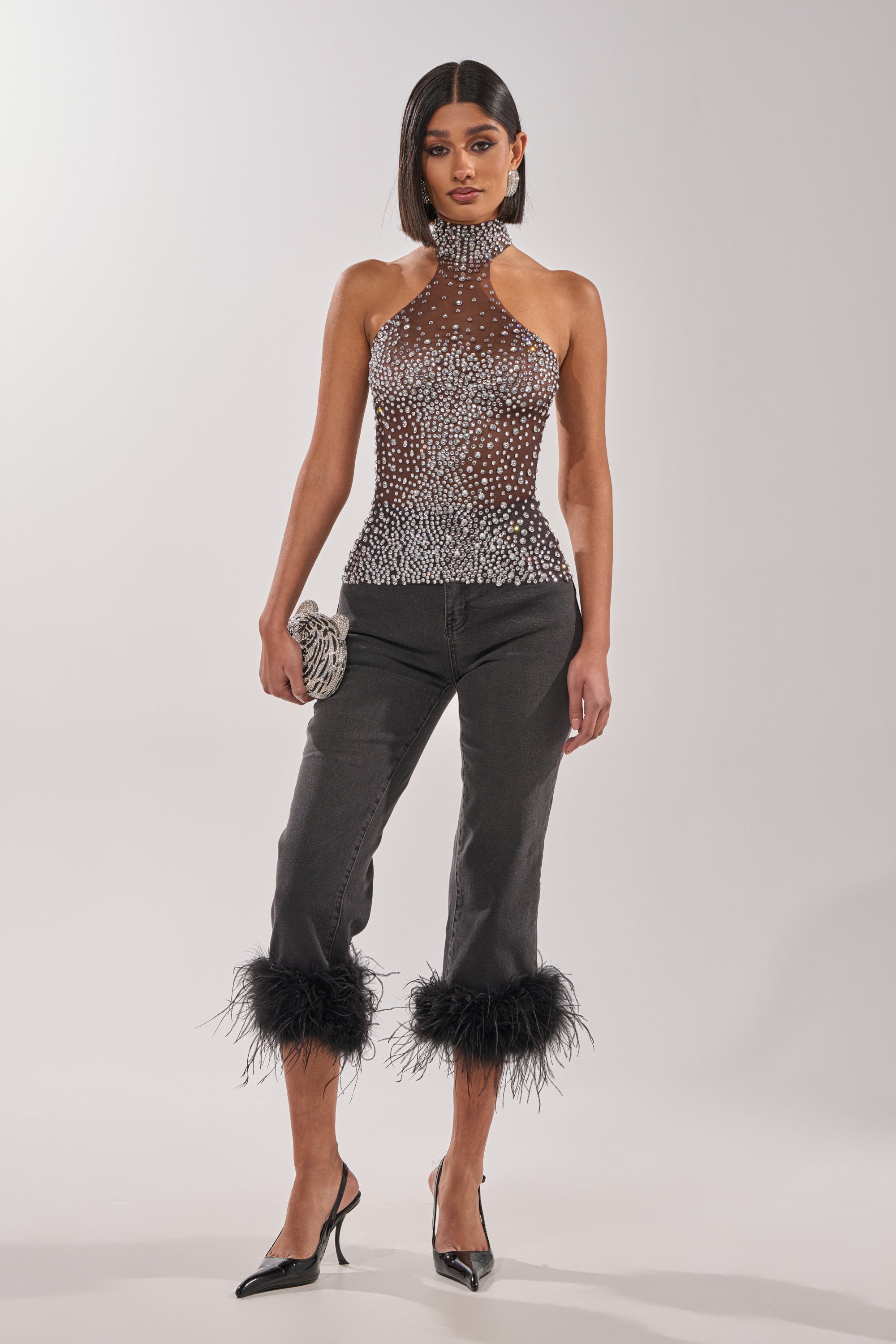A woman models the ALL YEAR ROUND SHEER RHINESTONE TOP, paired with black feather-hem cropped pants, black pointed heels, and a silver clutch against a plain light background. 4
