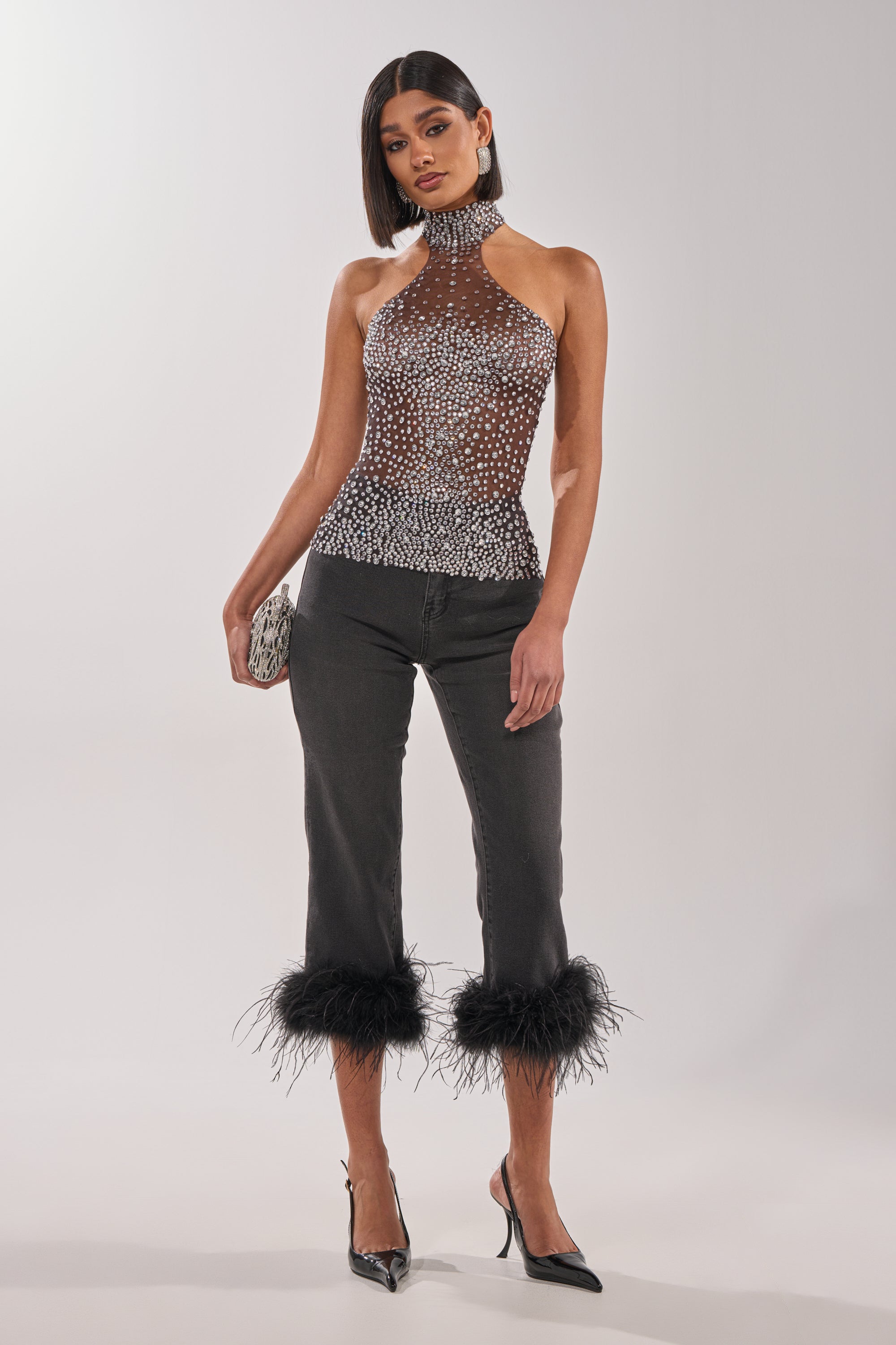 A woman poses against a plain white background wearing the ALL YEAR ROUND SHEER RHINESTONE TOP, cropped black pants with feathered hems, black pointed heels, and holding a silver clutch. 5