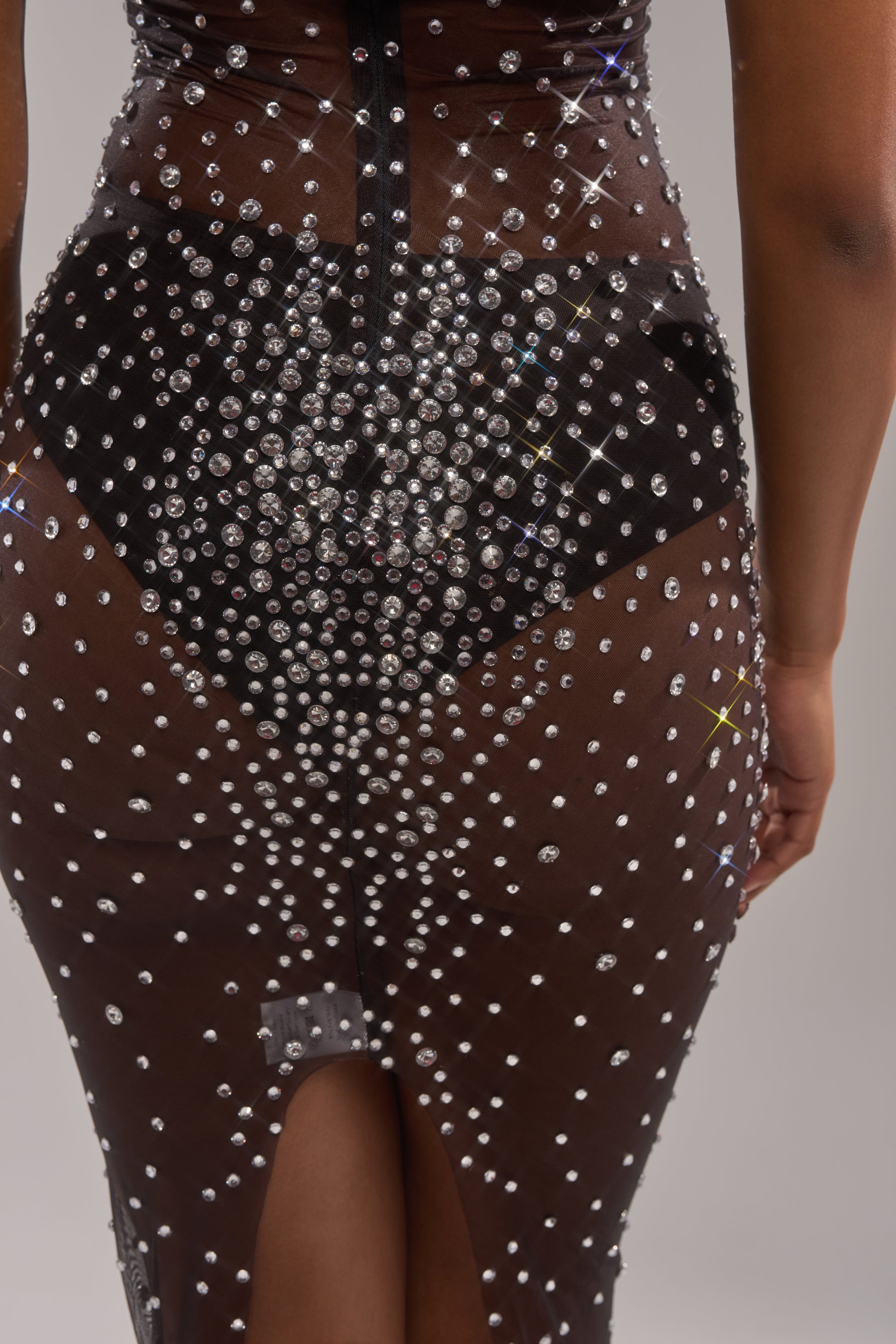 A close-up of the CRYSTAL CORE MAXI DRESS highlights its sheer black rhinestone mesh, sparkling crystal embellishments, visible black undergarments, central zipper, and a bottom slit for added detail.