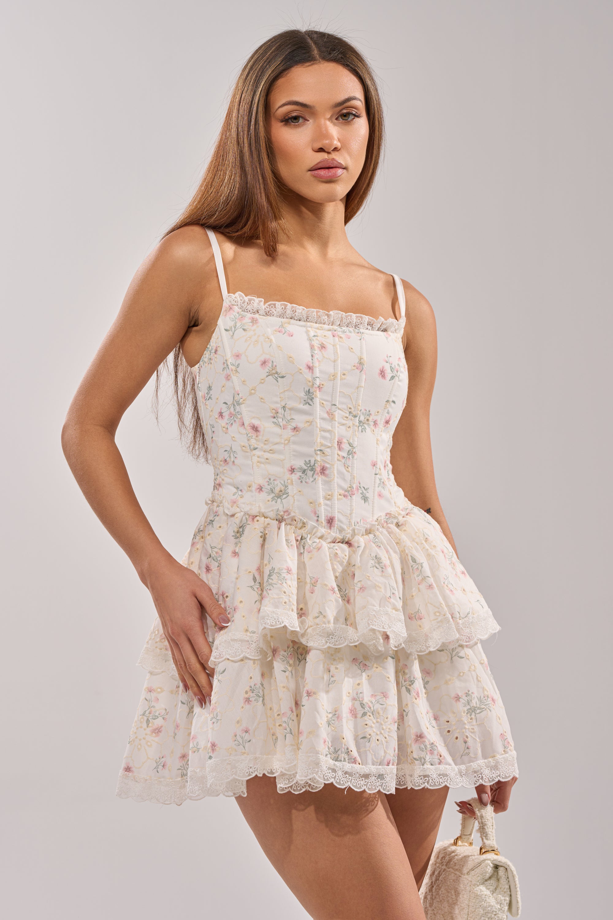 A woman in the TEA TIME MINI DRESS—a sleeveless, tiered, white floral corset mini dress with lace trim—stands against a plain light background, holding a small white textured handbag. 1