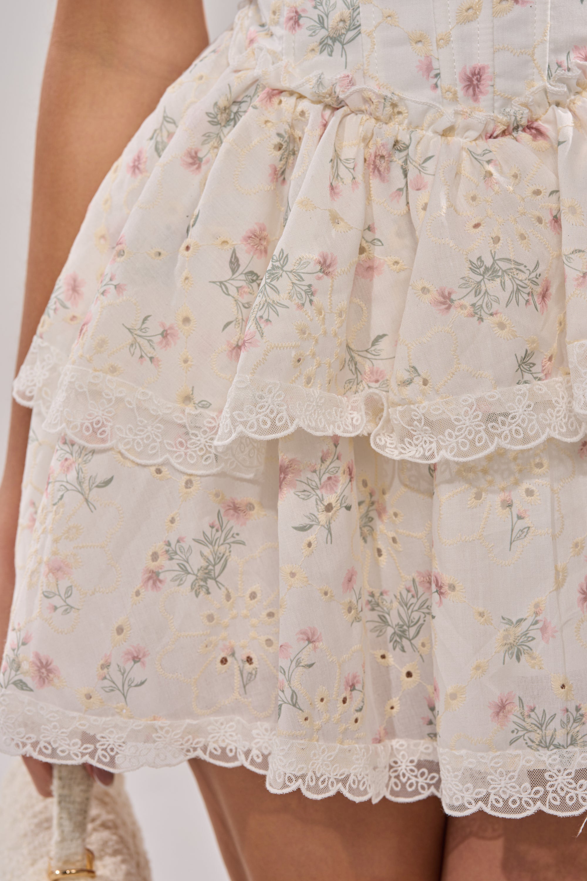 Close-up of the TEA TIME MINI DRESS featuring a white floral layered ruffled skirt with gold embellishments. Delicate lace trims the hem, and a hand holding a bag appears on the left. 7