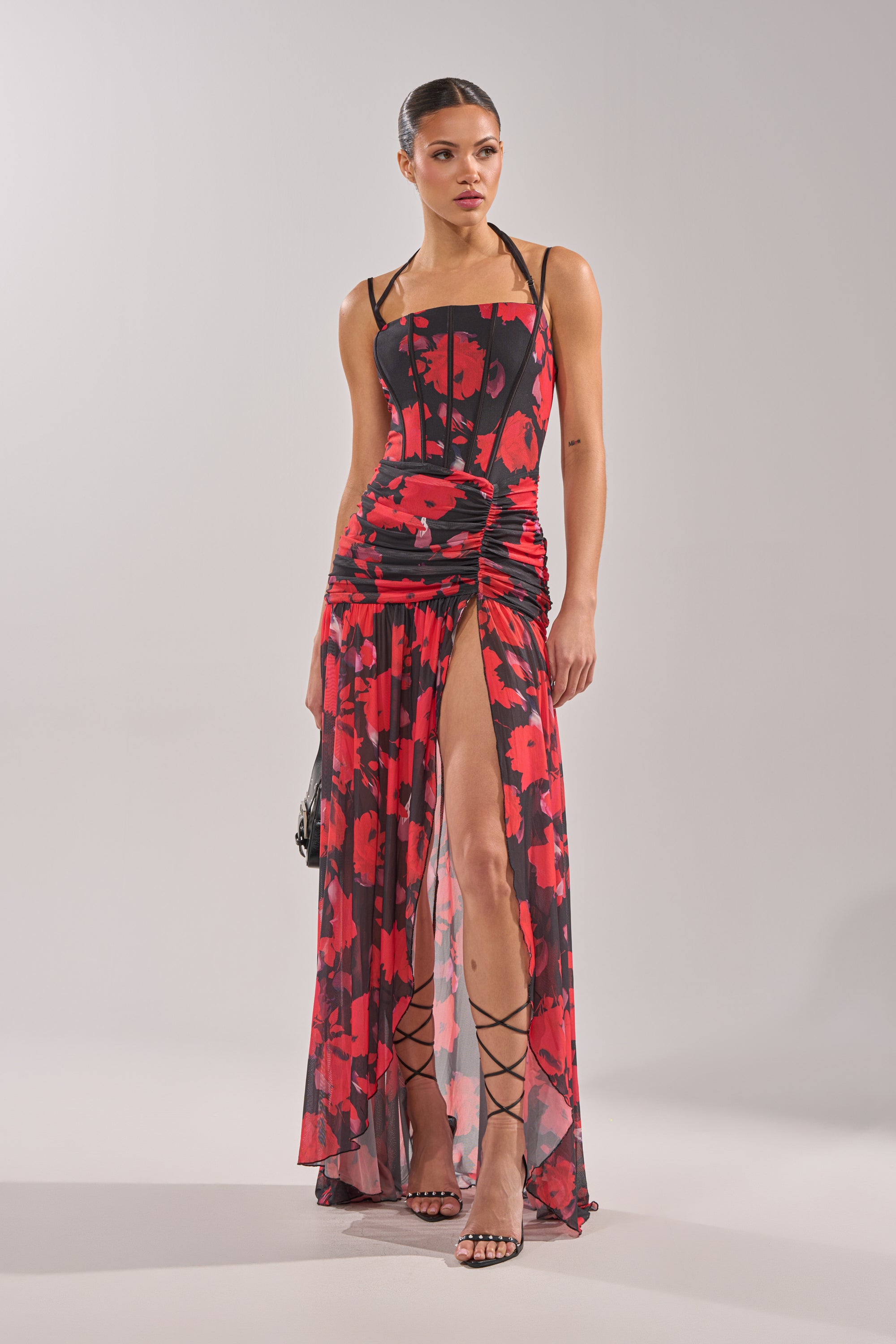 A woman poses against a light gray backdrop in the ROSE GARDEN MESH MAXI DRESS, featuring a red and black abstract floral print, spaghetti straps, ruched details, a high slit, and corset-style bodice, styled with strappy heels.