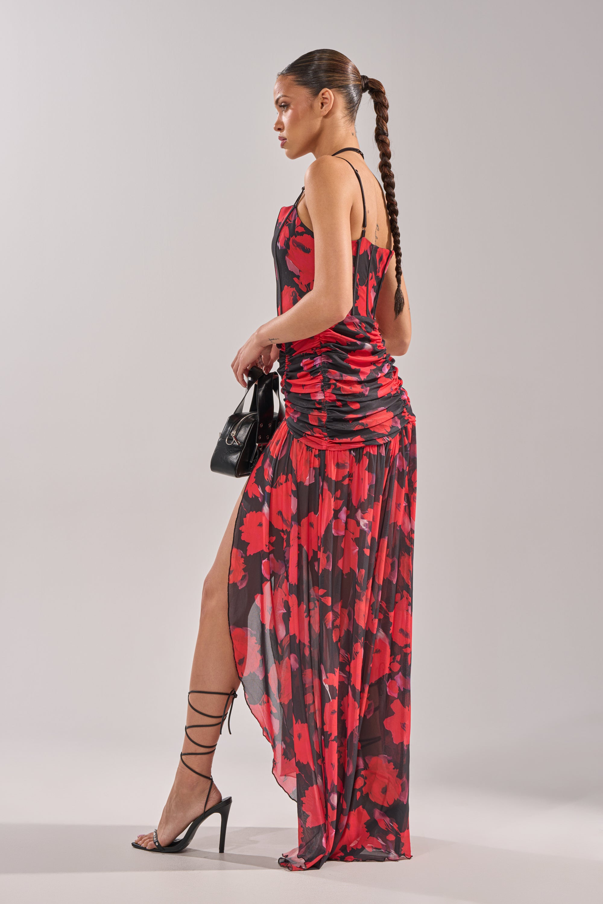 A woman stands sideways in the ROSE GARDEN MESH MAXI DRESS, a red and black abstract floral print design with spaghetti straps, a high slit, and asymmetrical hem. She has a long braid, strappy black heels, and carries a small black handbag.