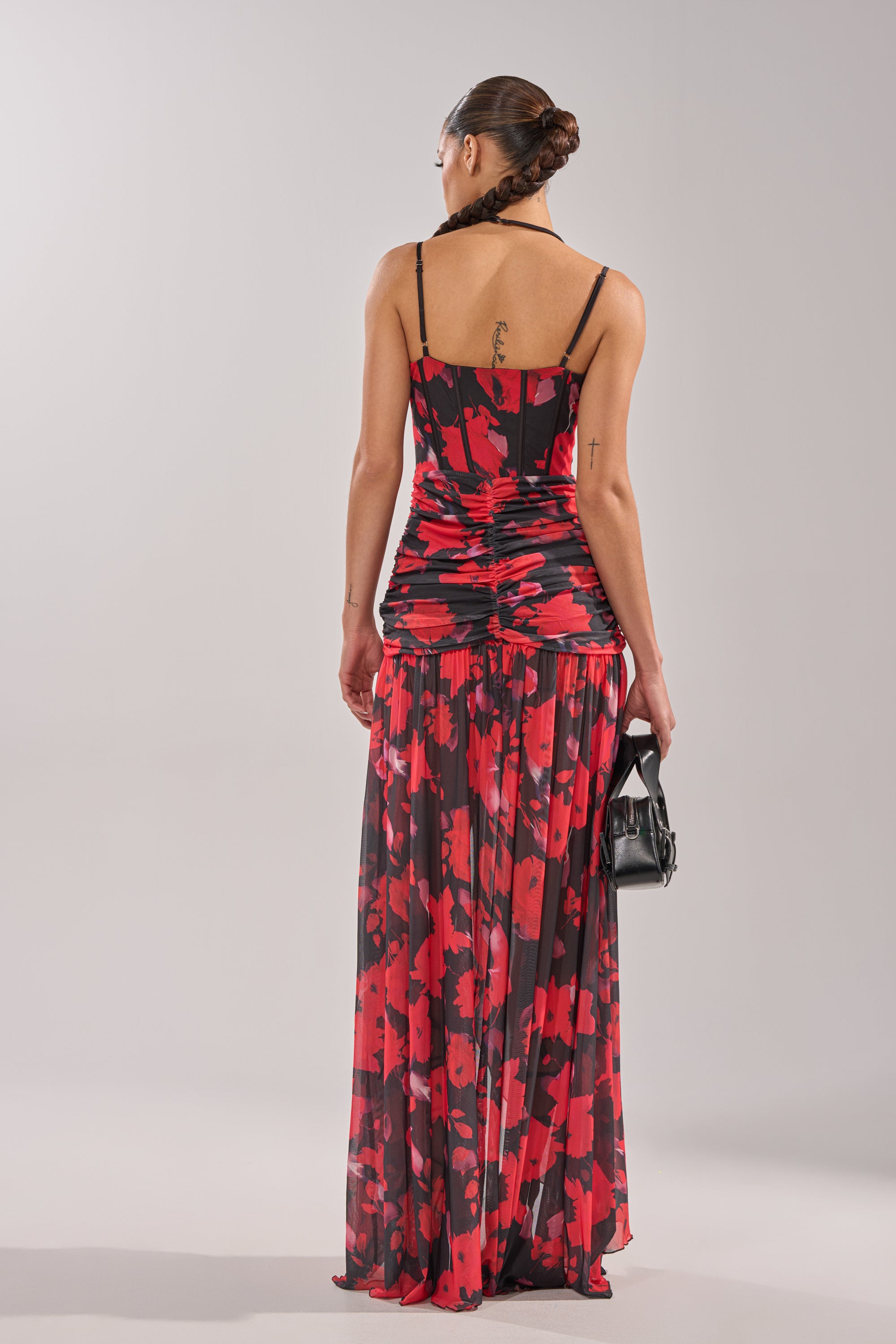 A woman with braided hair in an updo wears the ROSE GARDEN MESH MAXI DRESS, a long sleeveless floral mesh dress in red and black, standing with her back to the camera while holding a small black handbag.