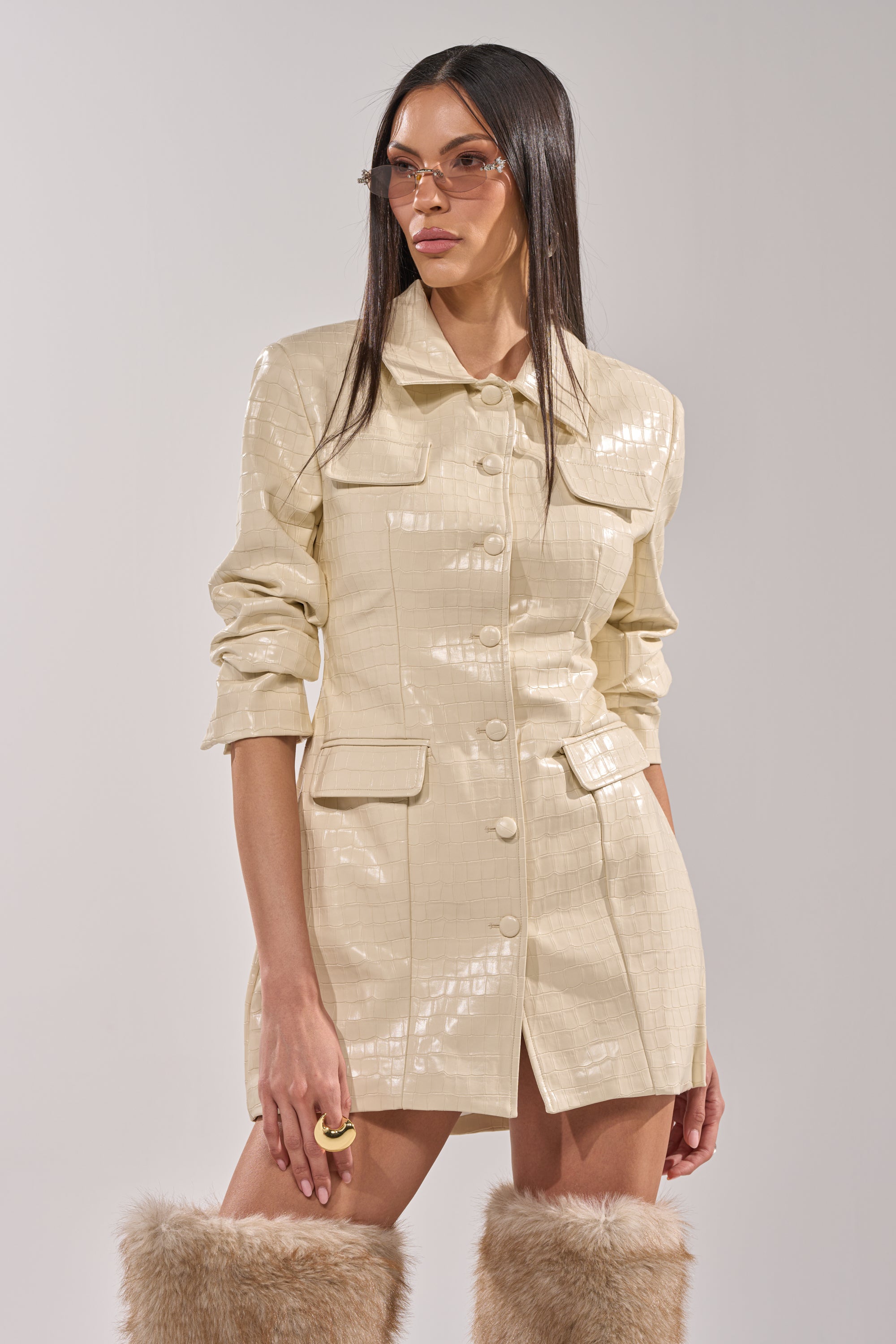 A woman wears the STANDING ON BUSINESS PU MINI DRESS, an ivory mini with front pockets, paired with beige sunglasses and tan furry knee-high boots, standing against a plain light background. 0