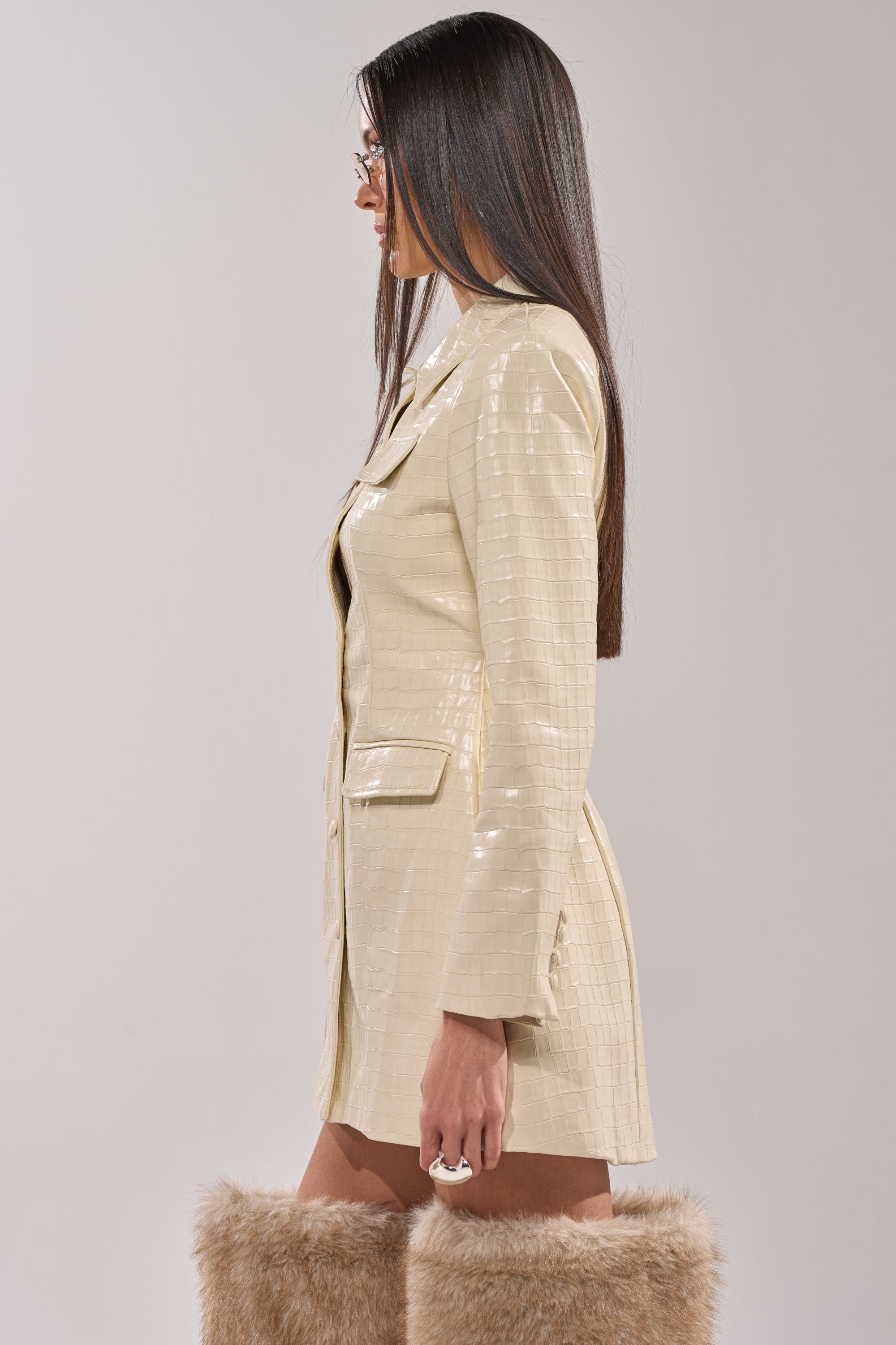 A woman with long straight hair models the STANDING ON BUSINESS PU MINI DRESS, a shiny light beige crocodile-embossed style, paired with tan faux fur knee-high boots, posing in profile against a plain background. 5