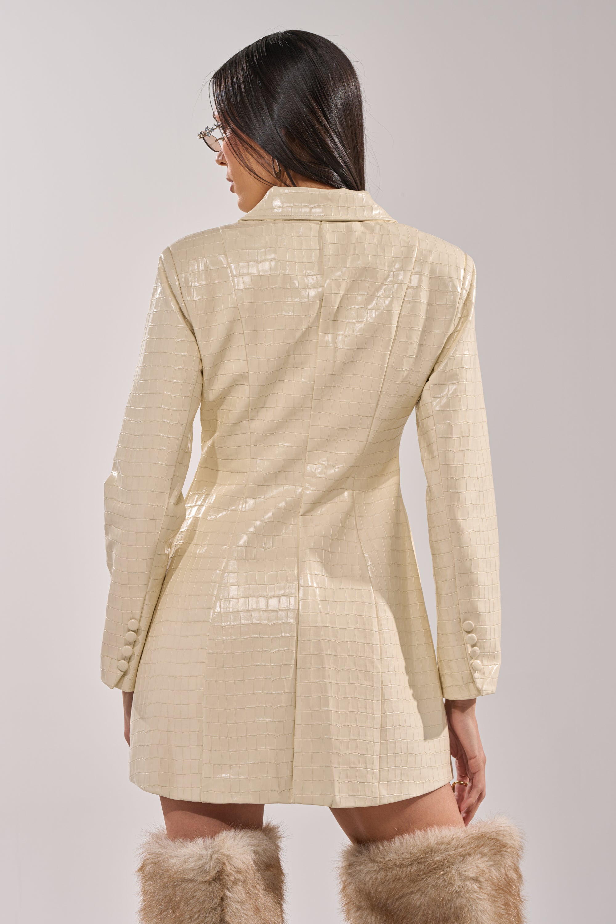 A woman with long dark hair and glasses is seen from the back, wearing the STANDING ON BUSINESS PU MINI DRESS in shiny ivory with textured details and knee-high furry beige boots, against a plain light background. 7