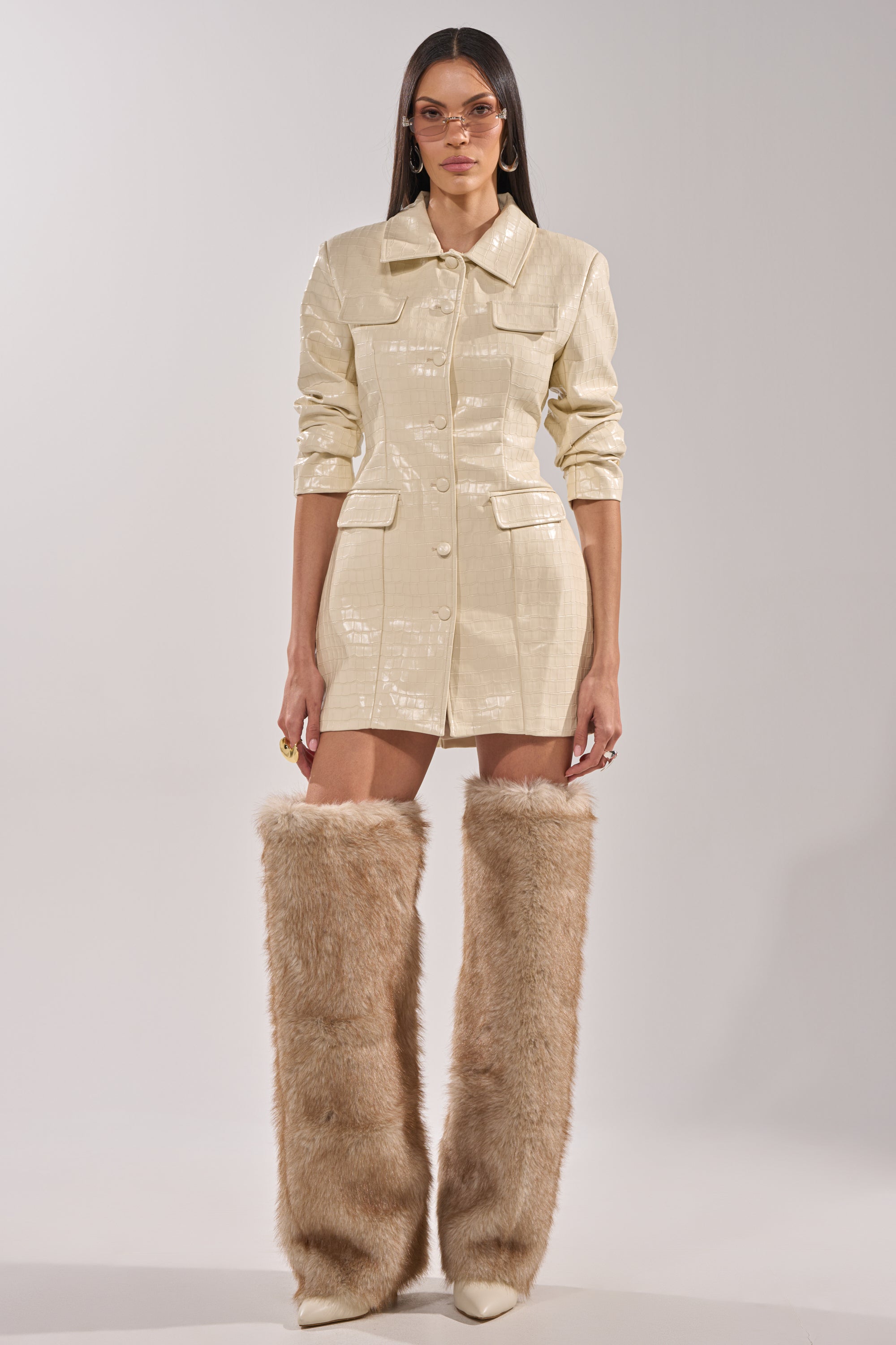 A woman poses against a plain backdrop wearing the STANDING ON BUSINESS PU MINI DRESS, styled as an ivory, shiny coat. She completes the look with oversized tan faux fur knee-high boots, sunglasses, hoop earrings, and long straight hair. 8