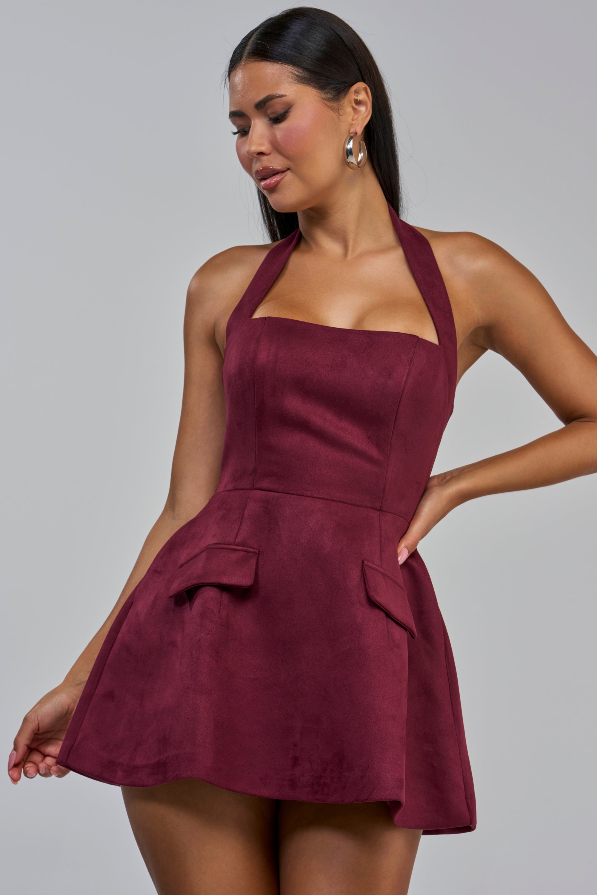A woman models the GET ME IN TROUBLE SUEDE MINI DRESS IN PLUM—a sleeveless, halter-style mini dress with front pocket flaps—against a plain gray backdrop. Crafted from chic faux suede in a rich plum shade, this dress offers a sophisticated yet playful look, making it the perfect autumn going out piece.