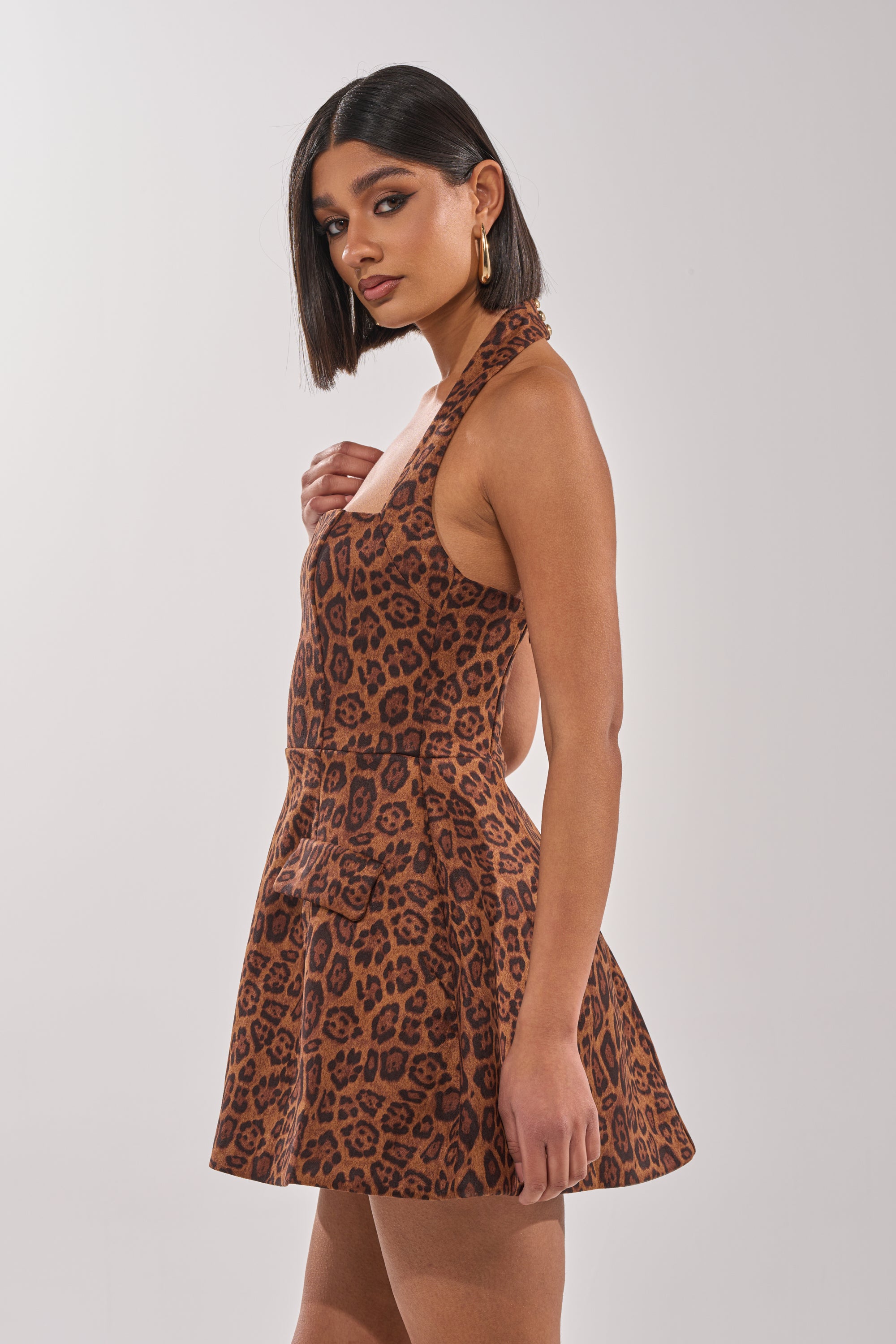 A woman with straight, shoulder-length dark hair wears gold hoop earrings and the CHEETAH GIRL MINI DRESS. She stands sideways against a plain light background, looking at the camera. 2
